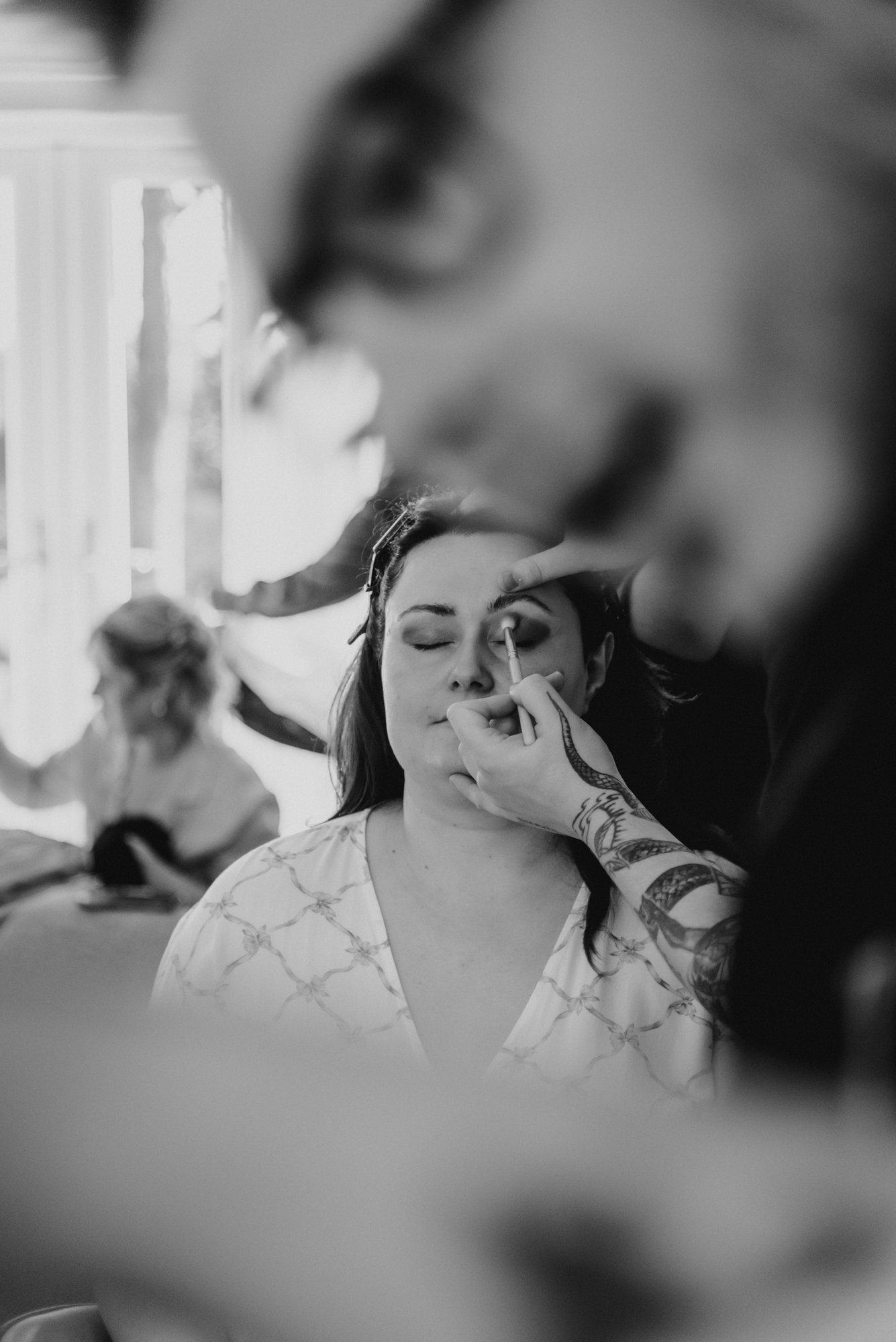 A woman with closed eyes getting her makeup applied by a makeup artist with tattoos on their arm, in black and white.