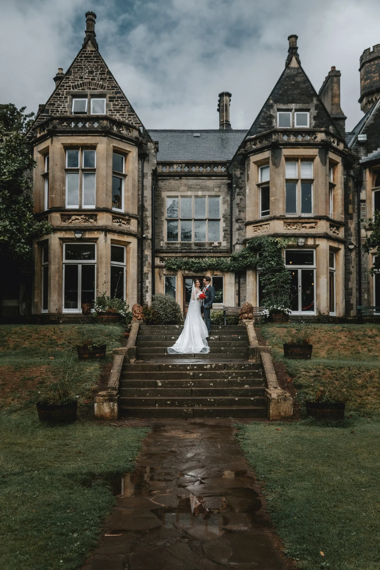 A bride and groom standing on the steps of a large, historic castle-like building with turrets, arched windows, and climbing plants, on a damp day.