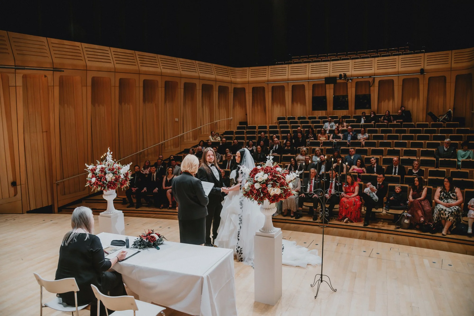 A wedding ceremony taking place in an auditorium with wooden walls. The bride and groom stand at the altar, exchanging vows, with an officiant and a woman seated at a table nearby. Two large floral arrangements are on either side of the altar, and gu