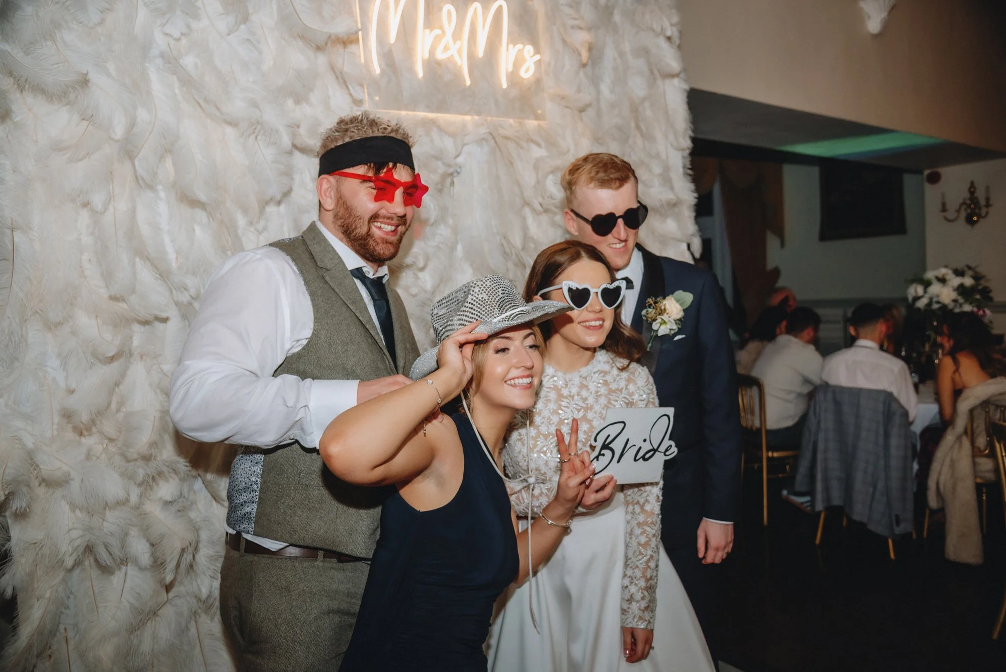 Group of four people at a wedding reception, wearing fun sunglasses and hats, smiling and posing for a photo in front of a white feathered backdrop with a neon sign reading 'Mr & Mrs'. One woman is holding a sign that says 'Bride' and making a peace 