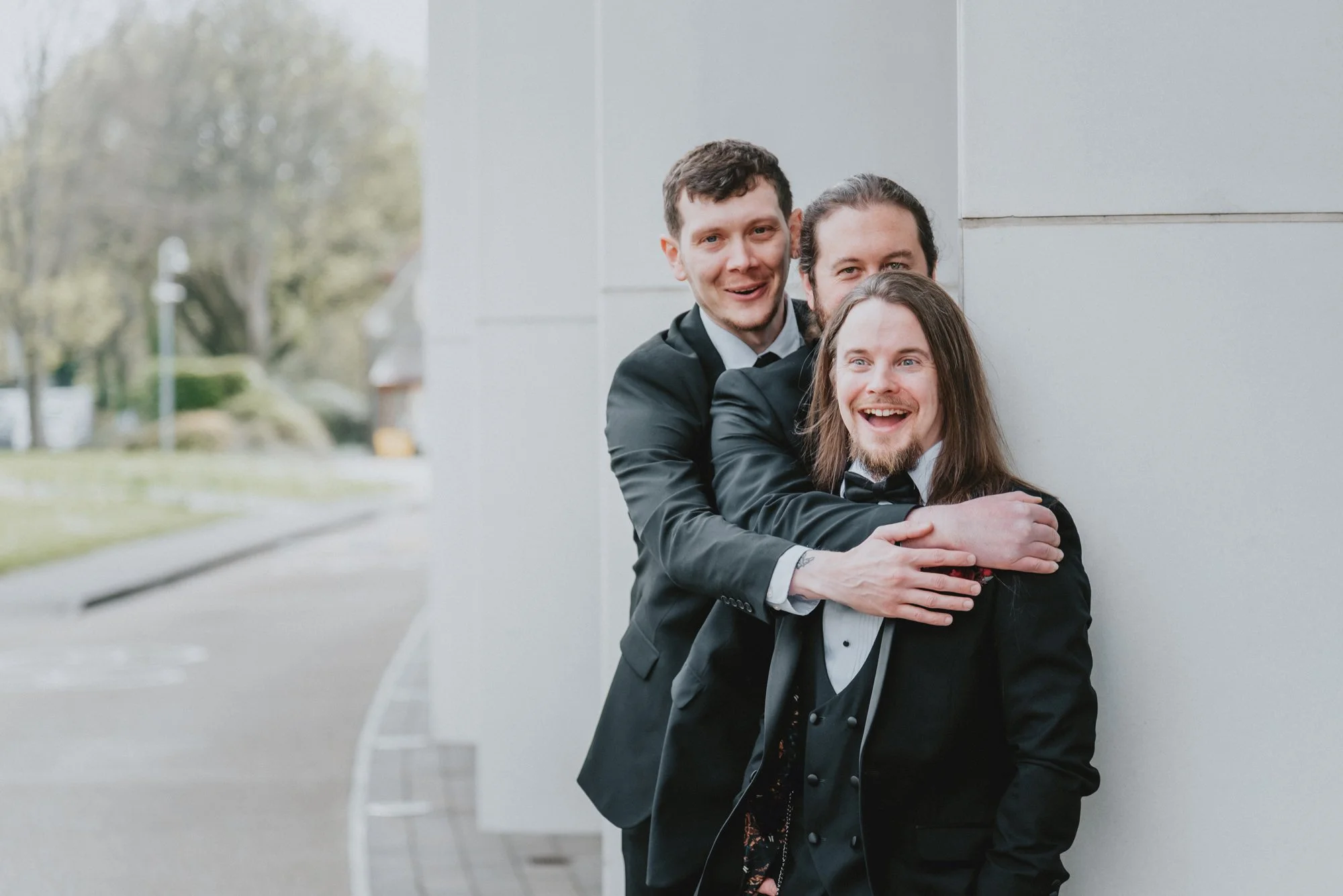 Three men in tuxedos enjoying a moment together outside, standing against a gray wall with a blurred outdoor background.