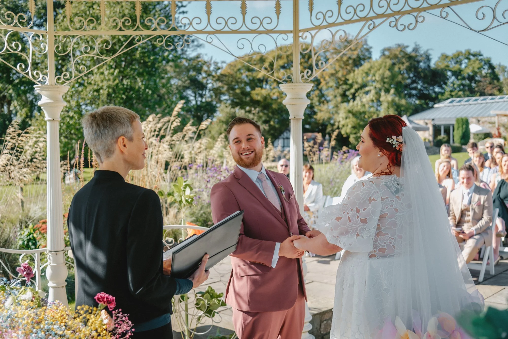 A wedding ceremony outdoors with the couple exchanging vows, surrounded by guests seated under a decorative white gazebo with a garden backdrop.