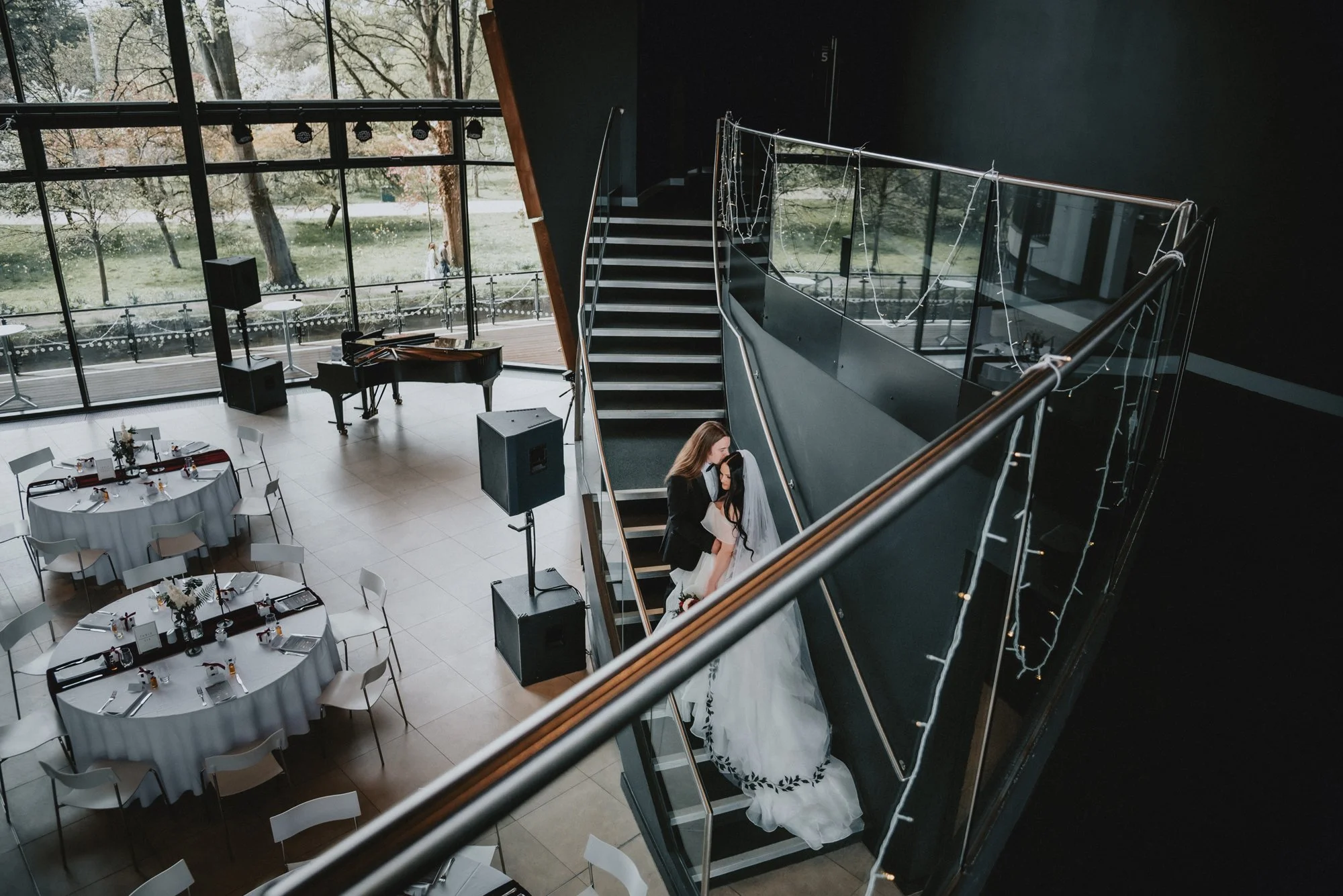 A bride and groom are on a staircase, sharing a kiss at a wedding reception with round tables set with white tablecloths and place settings, a grand piano, and large windows showing an outdoor view.