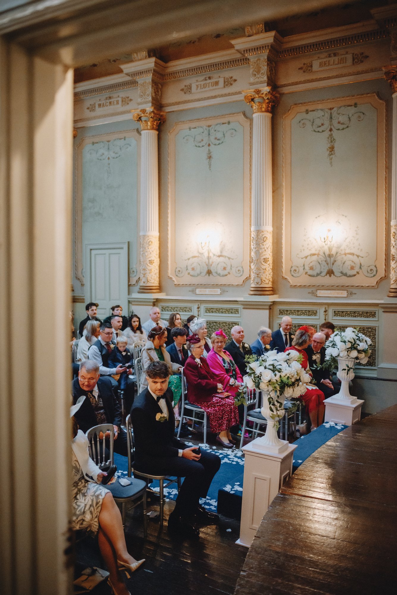 People attending a wedding ceremony in an ornate, classical-style room with cream and gold accents, large floral arrangements, and classical columns.
