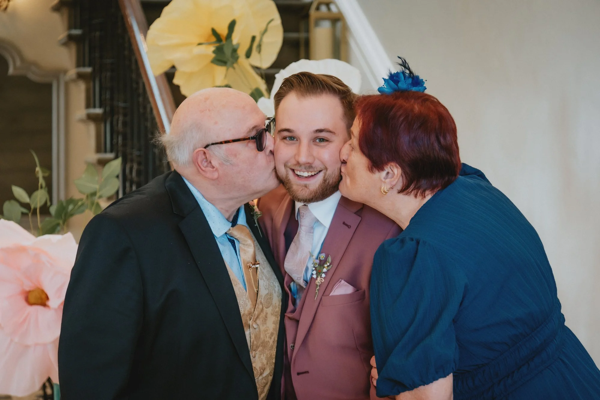 A young man in a pink suit being kissed on the cheeks by an elderly man and woman