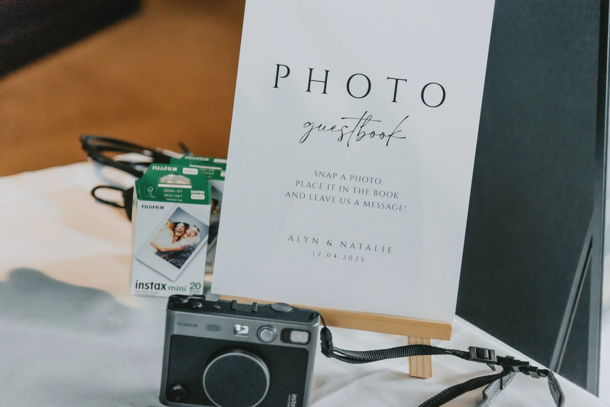 Photo guestbook sign, Fujifilm Instax Mini camera, and film on a table, with a black speaker in the background.