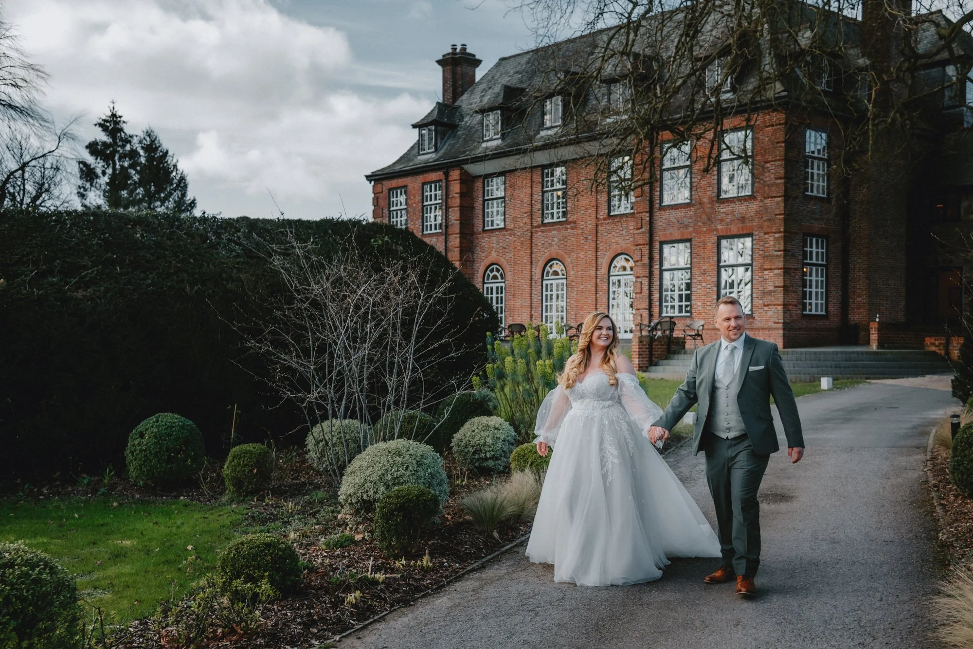 A newly married couple walking hand-in-hand on a driveway near a large brick house with numerous windows, surrounded by landscaped bushes and trees.