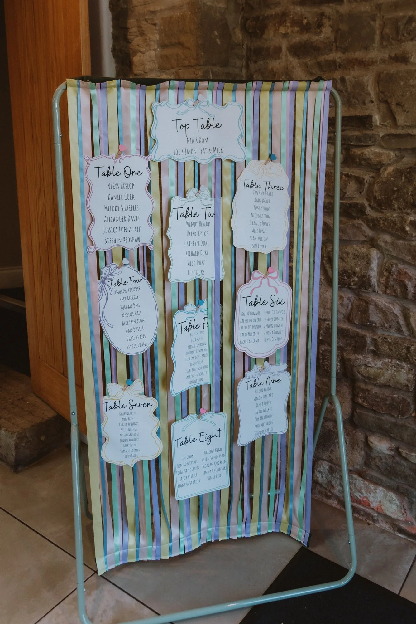 Wedding seating chart on a striped fabric backdrop with handwritten style labels for each table, listing guest names.