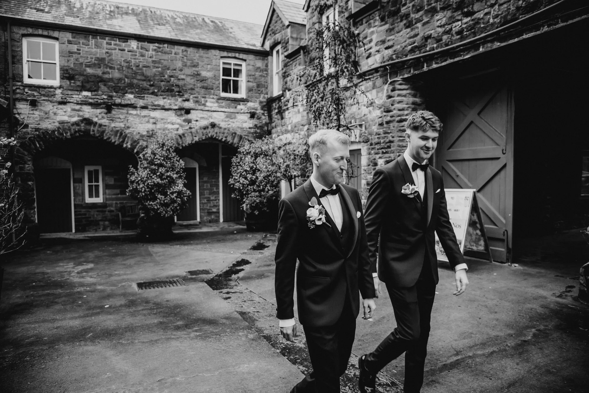 Two men in suits and bow ties walking together outside a stone building with arched doorways and greenery.