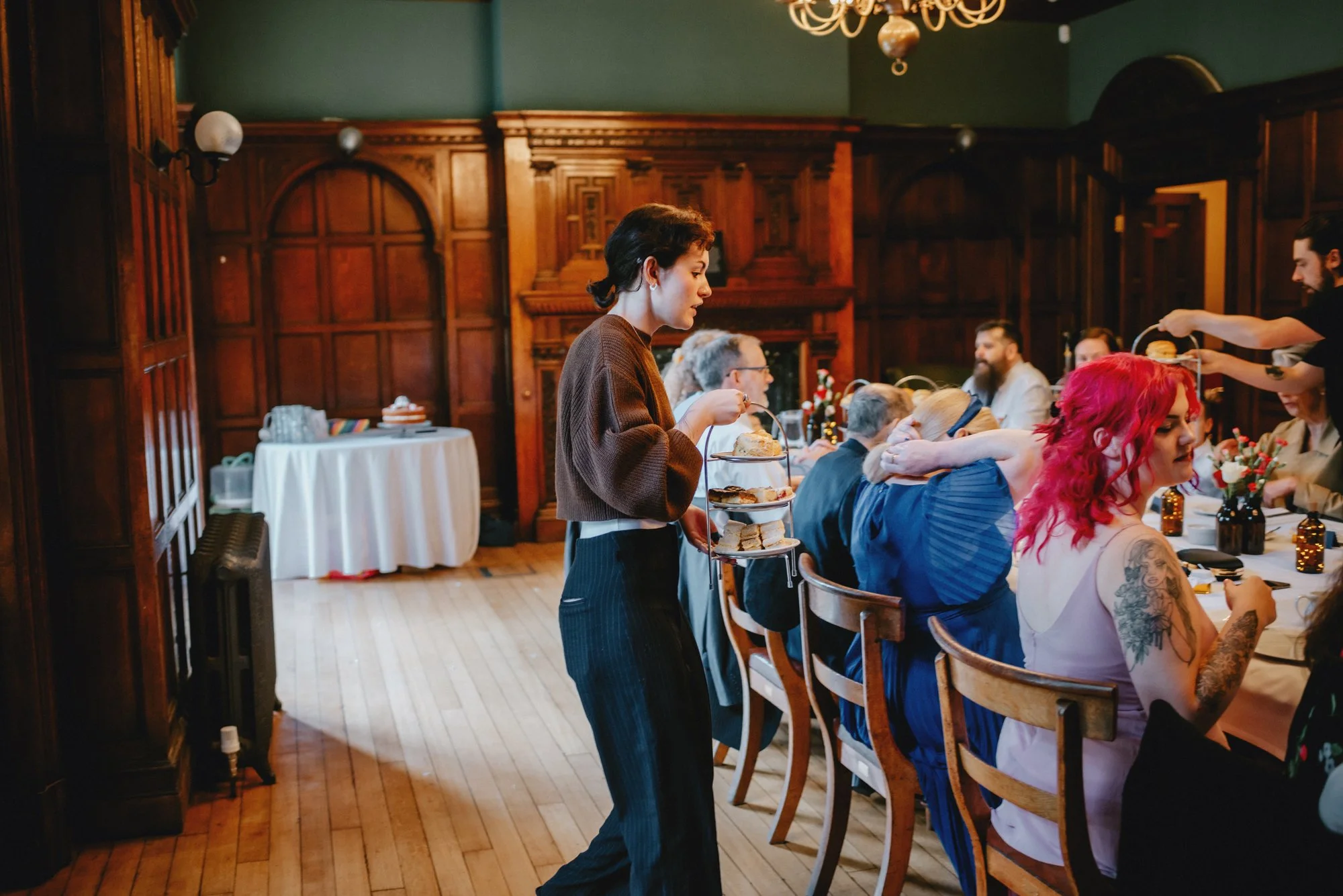 People sitting at a long table in a wooden-paneled room with a woman serving food with a tiered tray.