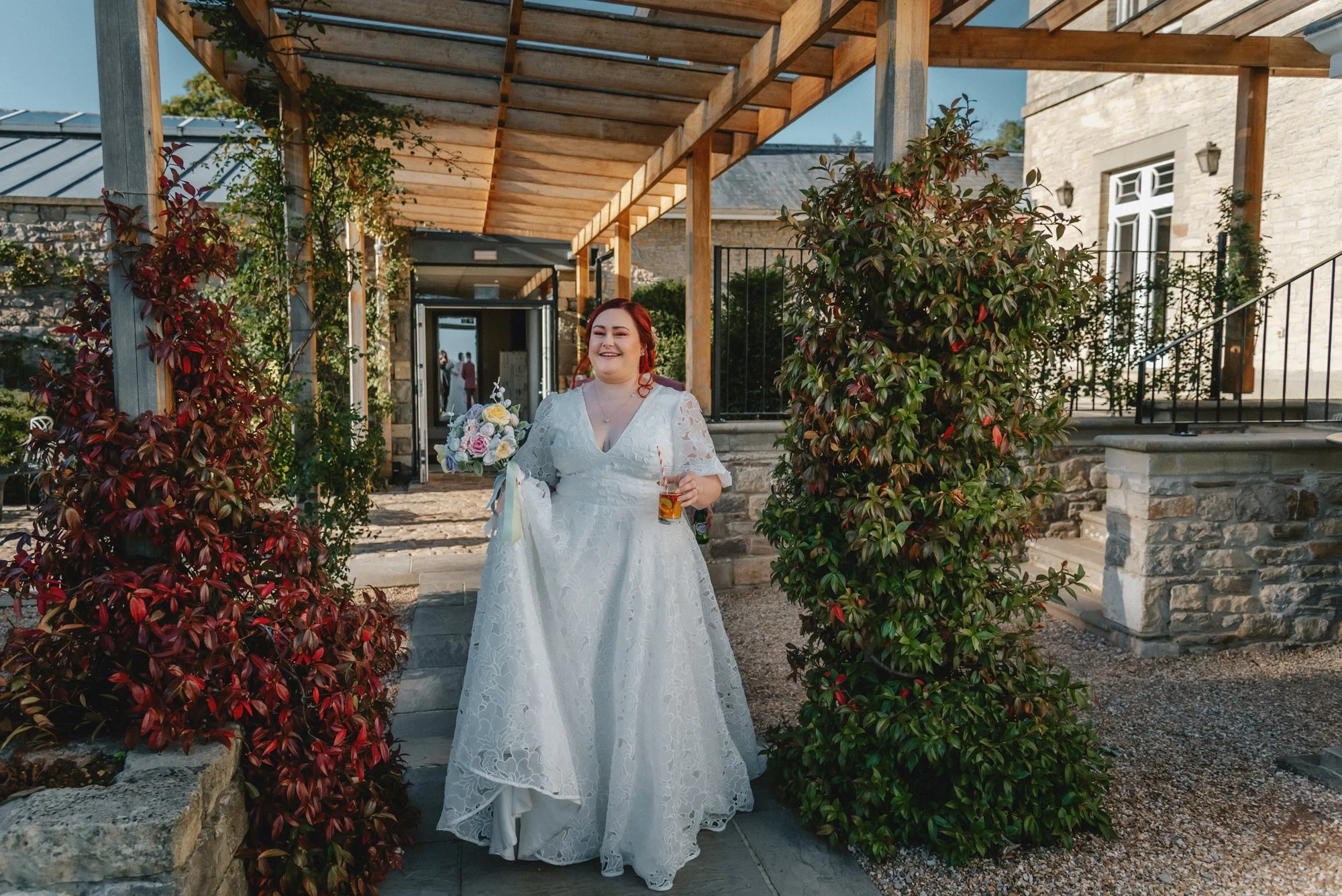A woman in a white lace wedding dress holding a bouquet and a drink, smiling as she walks through an outdoor wedding venue with stone and wood structures and green shrubbery.