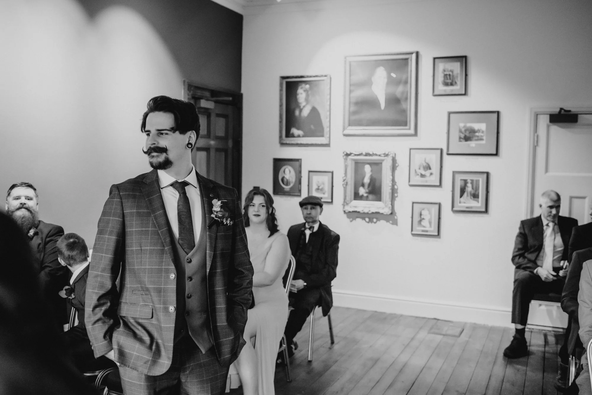 Black and white photo of a young man with styled hair and a beard, wearing a checkered suit, standing in a room with multiple framed paintings and photographs on the wall. Several other people are seated in the background, some looking at the young m
