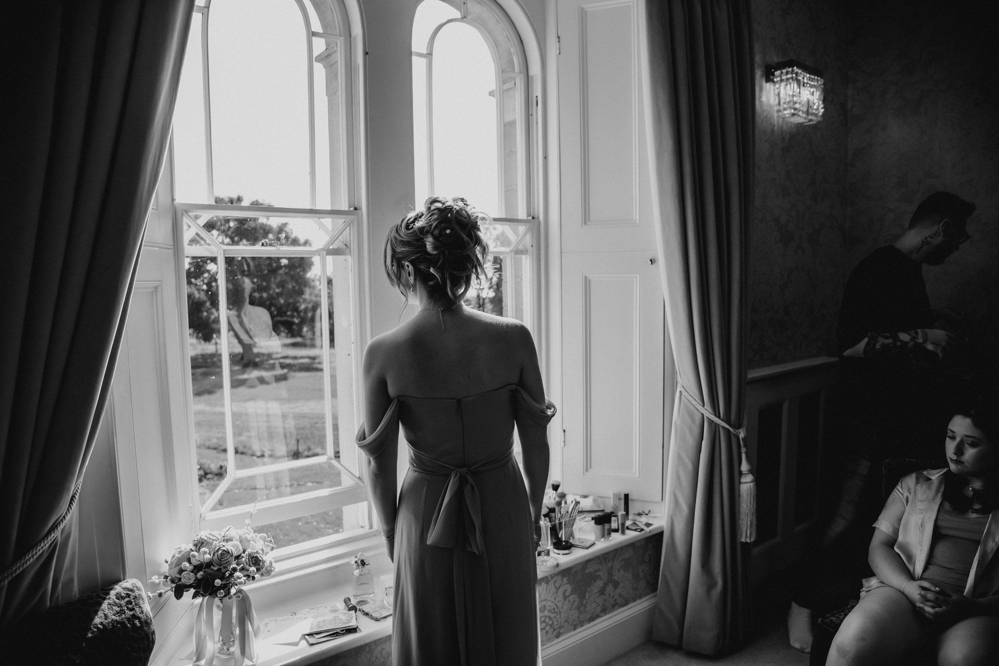 A woman with styled hair in a strapless dress stands near an open window, looking outside in a well-lit room. The room has elegant curtains, a bouquet of flowers, and various personal items on a windowsill, with two women sitting and a man standing i