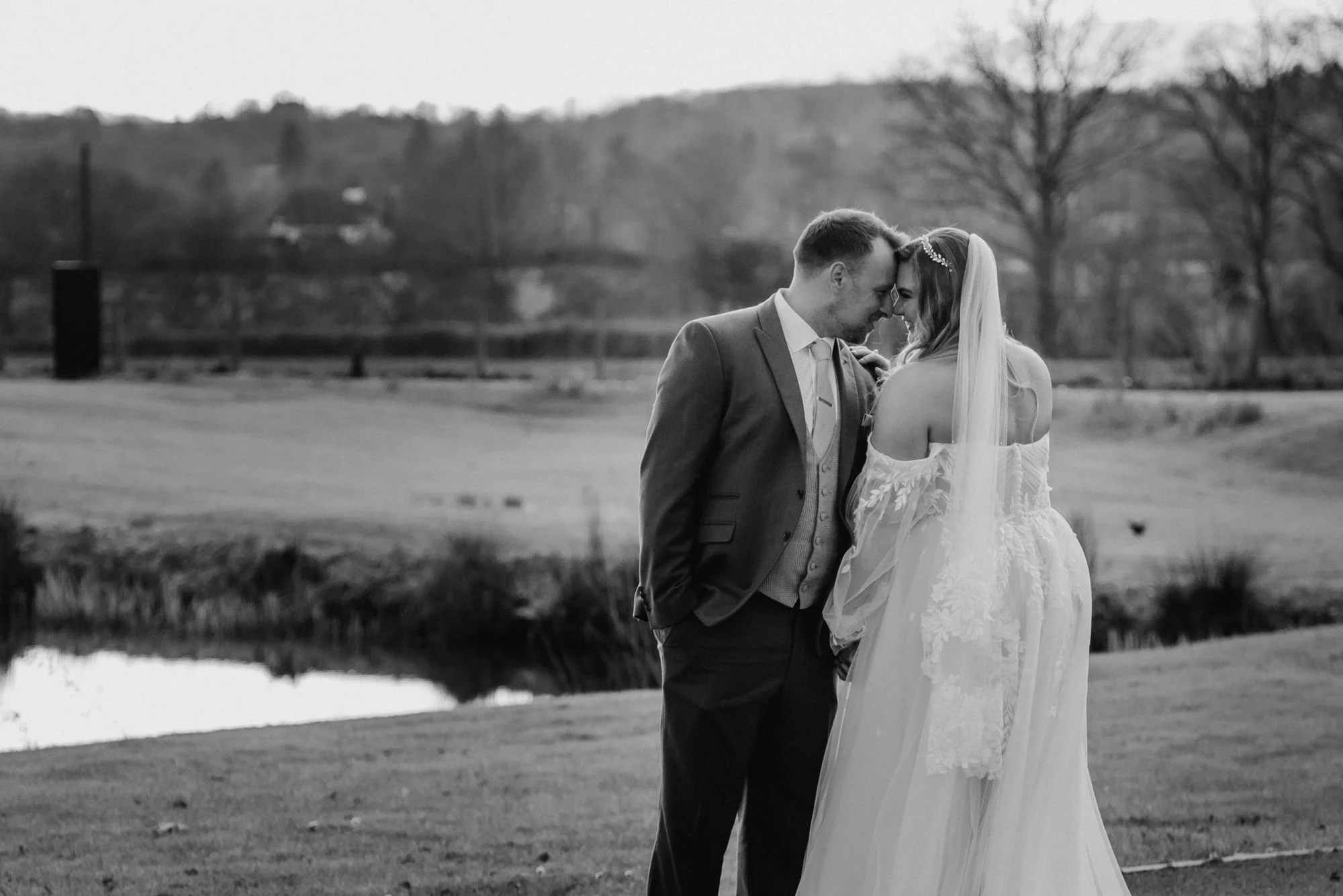 Black and white photo of a bride and groom touching foreheads outdoors, with trees and rolling hills in the background.