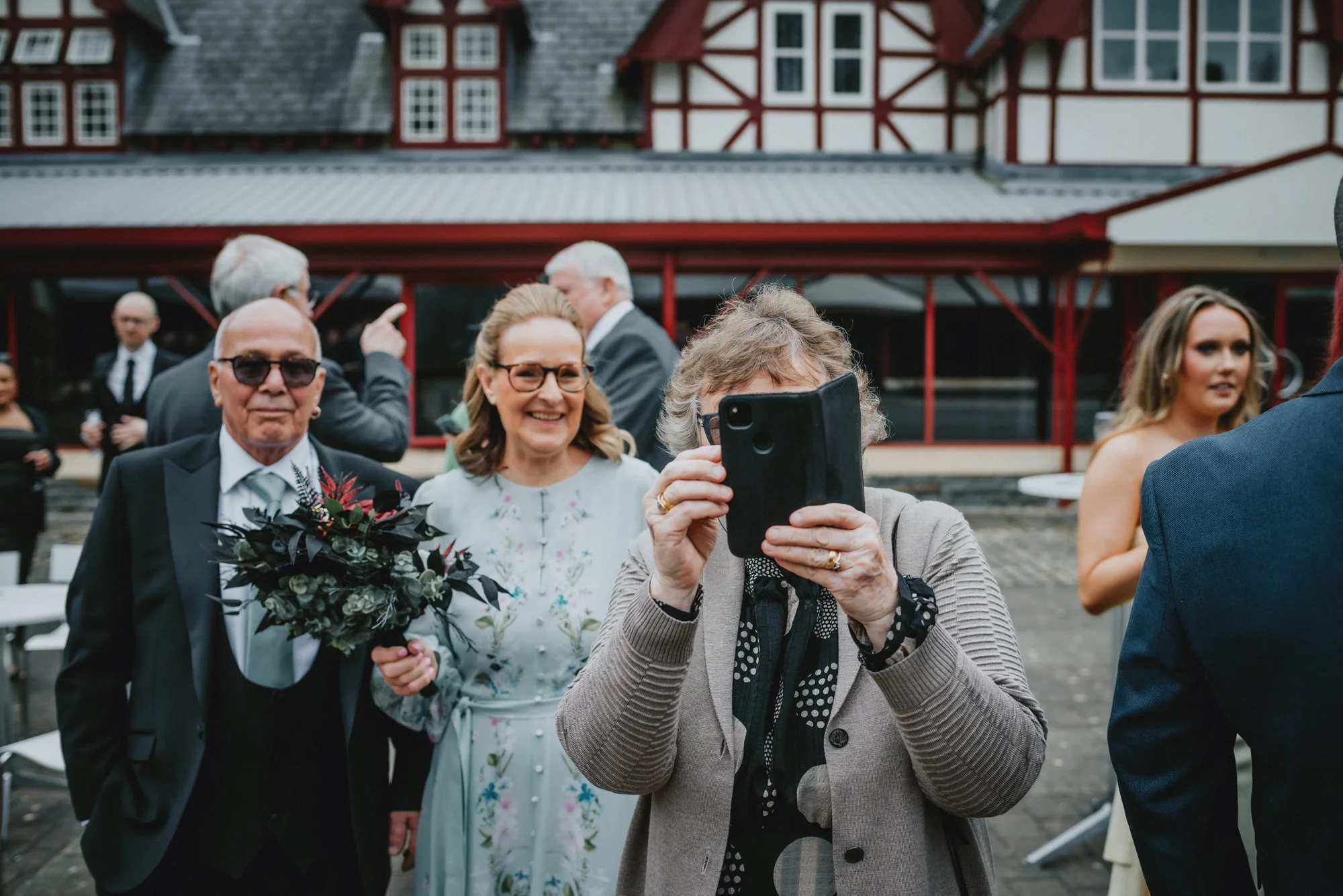 People at a wedding or social event, with one person taking a photo of others, including an elderly man holding a bouquet and a woman in a floral dress, outdoors in front of a building with a red and white exterior.