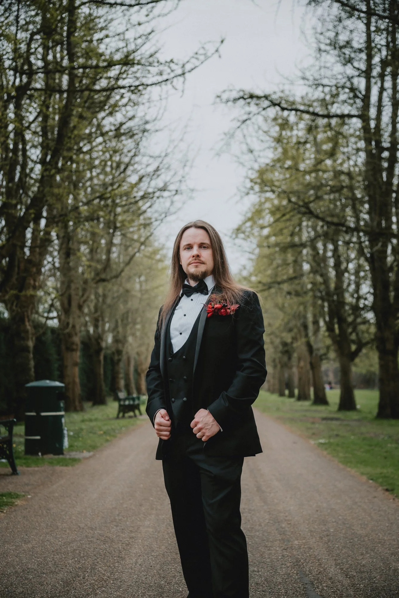 A man in a black tuxedo with a white shirt and black bow tie, standing on a tree-lined pathway in a park during daytime, with a red flower boutonniere on his lapel.