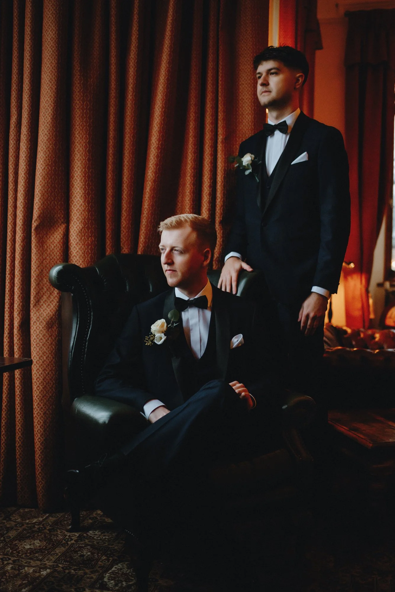 Two men in tuxedos with bow ties and white flowers on their lapels, one seated in a black leather armchair and the other standing behind him, inside a room with warm lighting and red curtains.