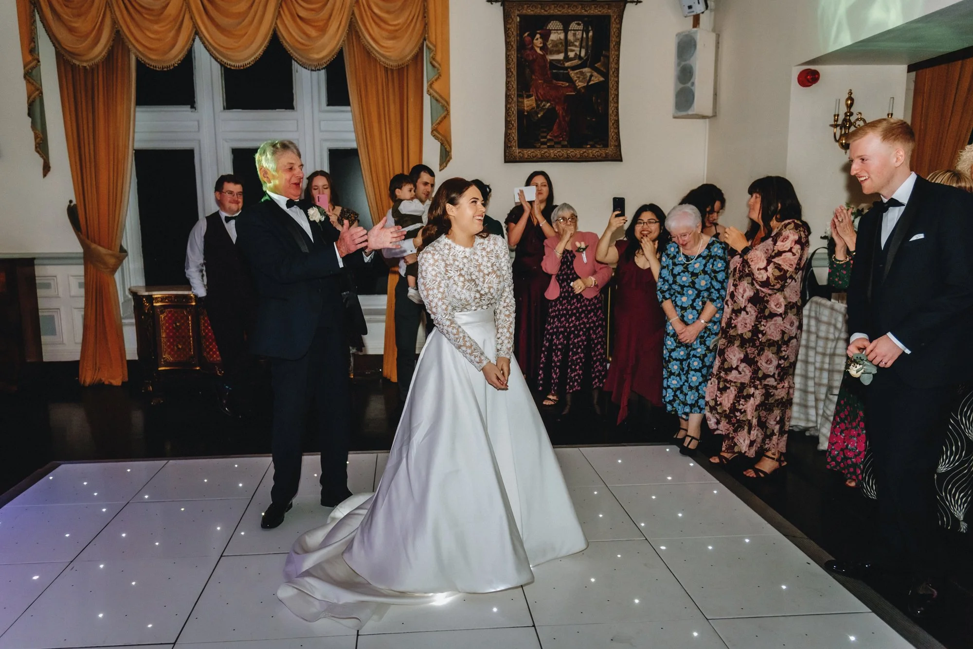 A bride in a white wedding dress with lace top stands on a lit dance floor, surrounded by guests at a wedding reception. An elderly man in a black tuxedo appears to be giving a speech or toast, while guests smile, clap, and take photos. The setting i