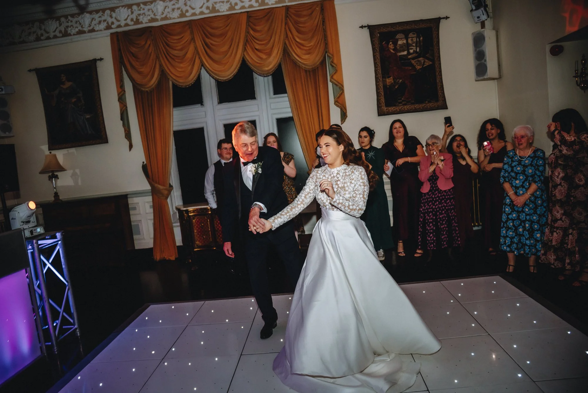 A bride and an older man dancing together at a wedding reception, surrounded by guests in a decorated hall.