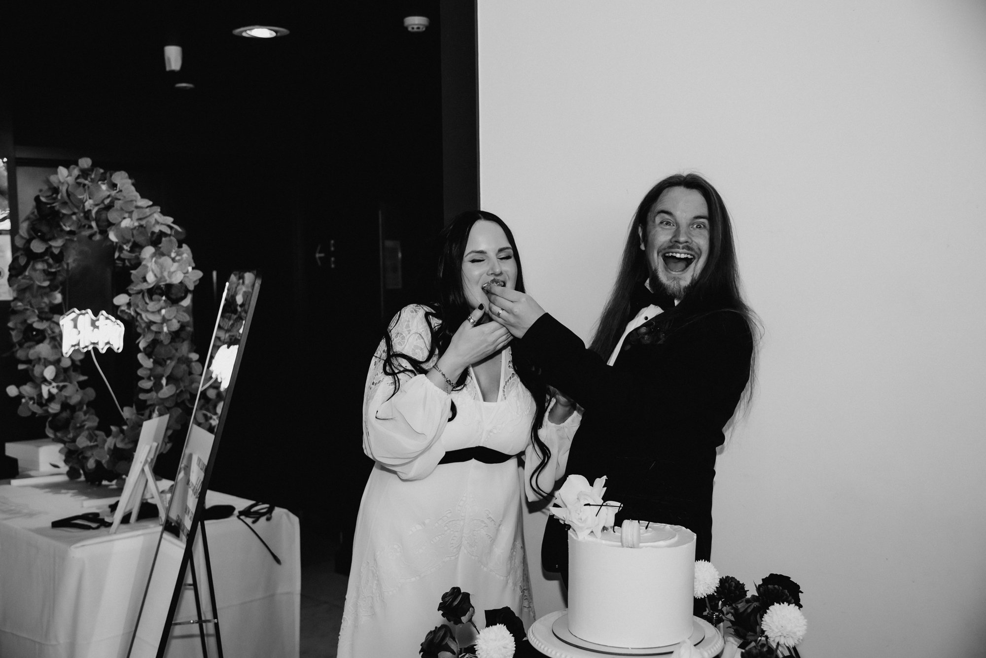 A happy couple celebrating at their wedding, feeding each other cake, with a wedding cake decorated with flowers on a table nearby.