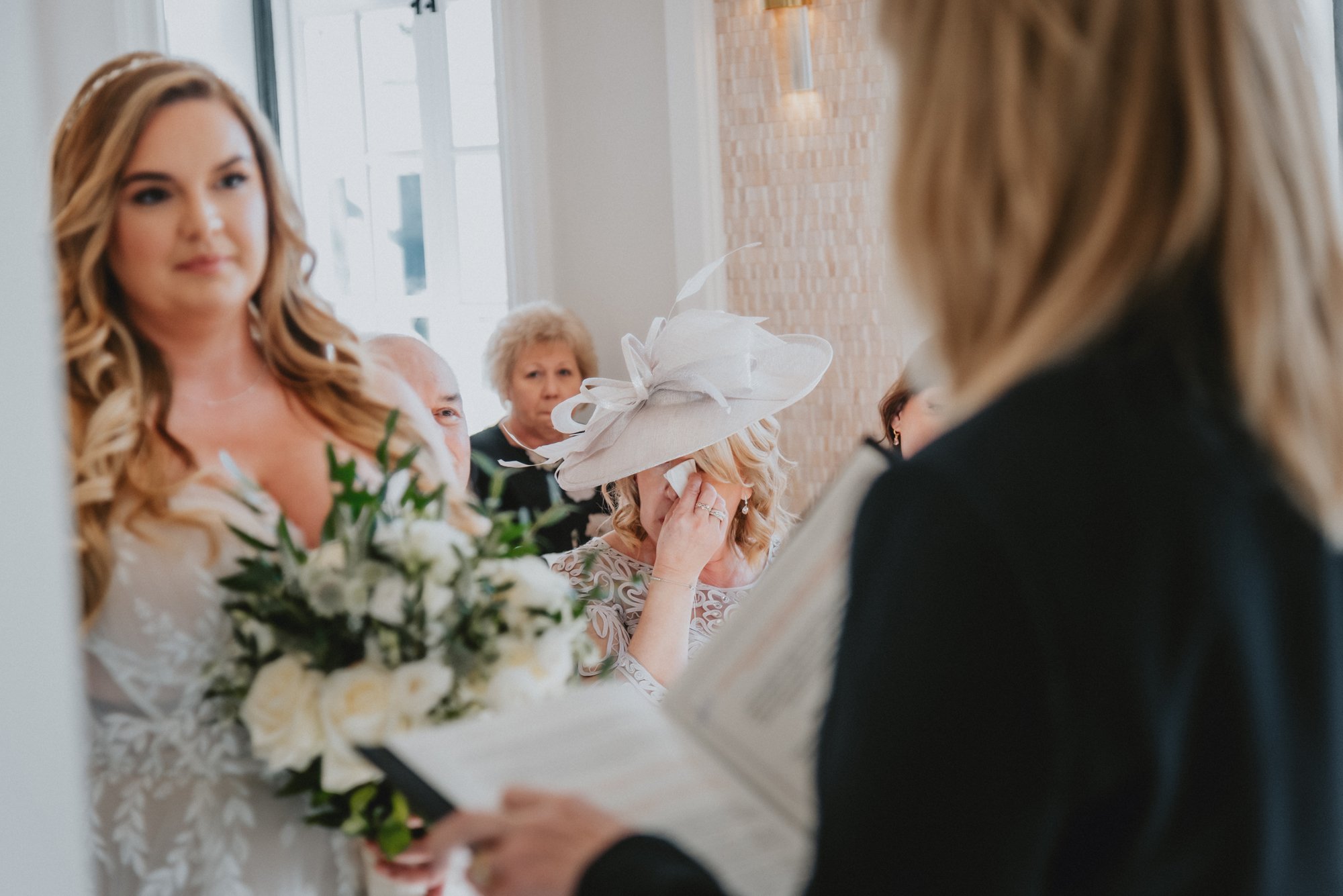 A bride with long wavy hair and a white wedding dress holding a bouquet of white flowers, facing an officiant during a wedding ceremony. An older woman wearing a large white hat with decorative feathers appears emotional, wiping away tears, in a room