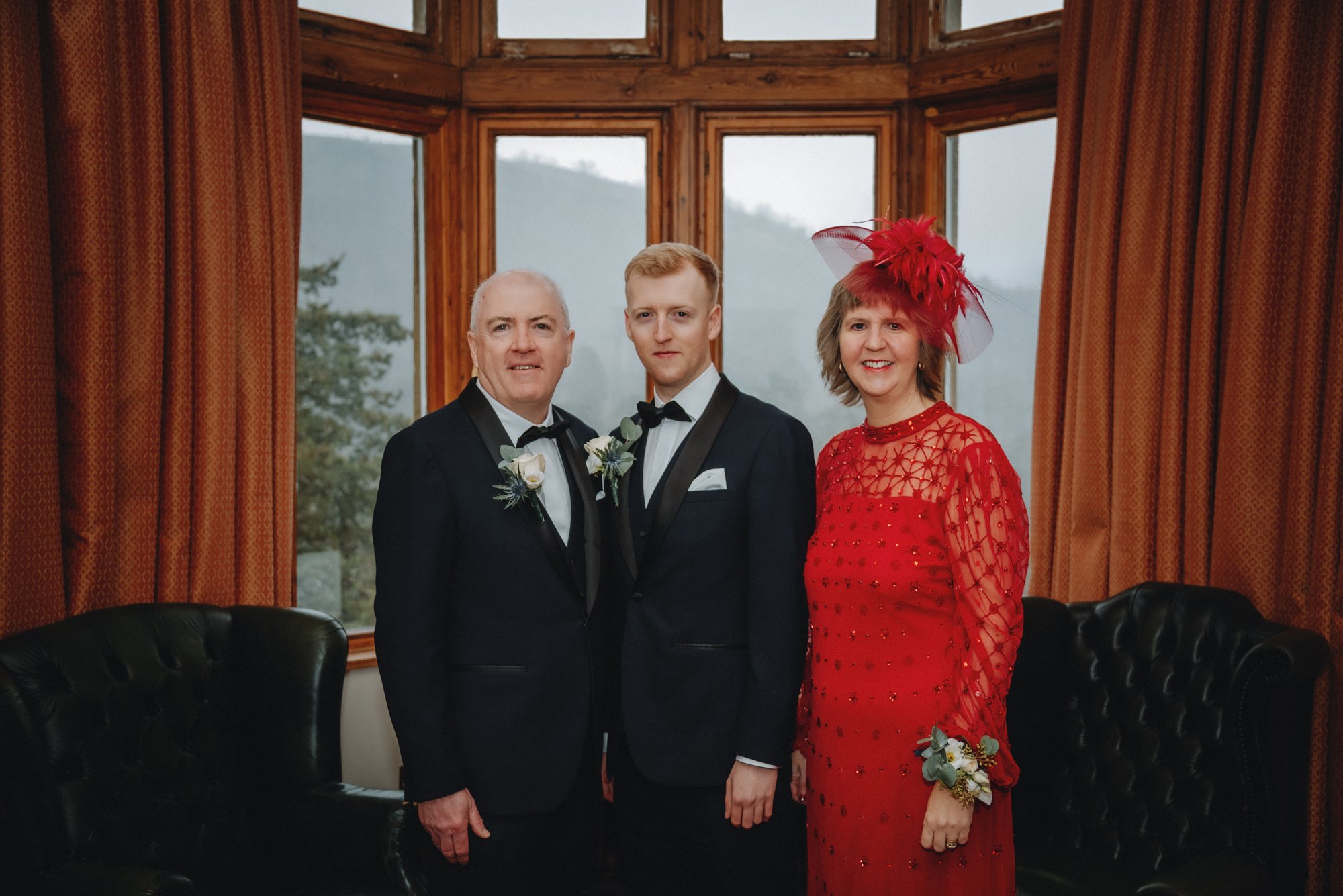 Three people standing indoors in a wooden room with large windows. The man on the left is wearing a tuxedo with a boutonniere, the man in the middle is wearing a tuxedo with a bow tie, and the woman on the right is wearing a red dress with a matching