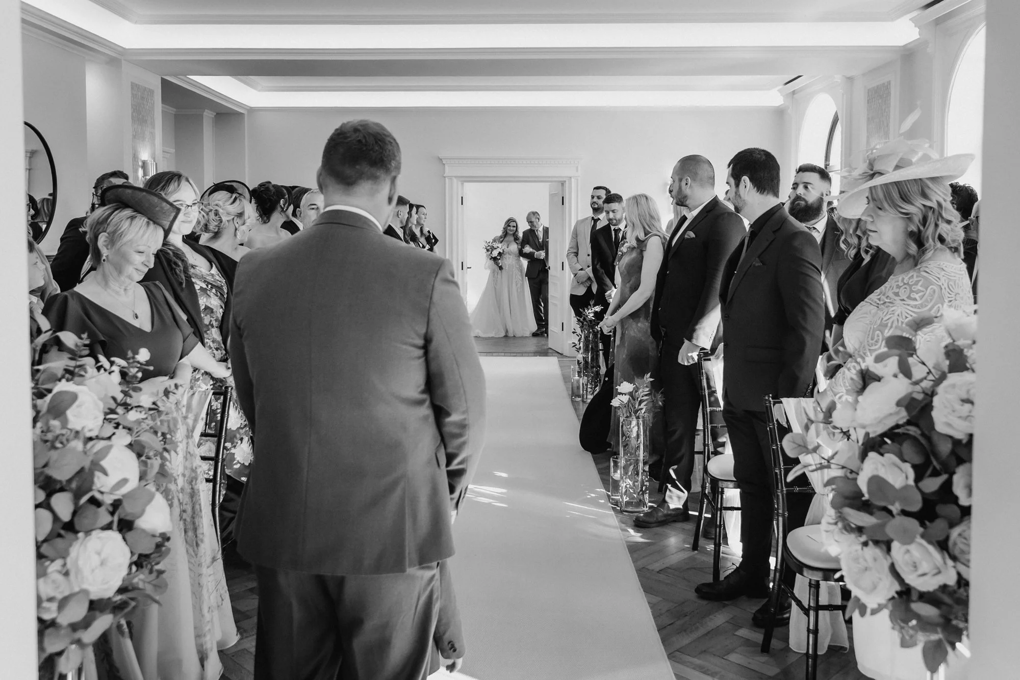 Black and white photo of a wedding ceremony with guests standing on both sides of an aisle, and the bride walking down the aisle towards the groom.