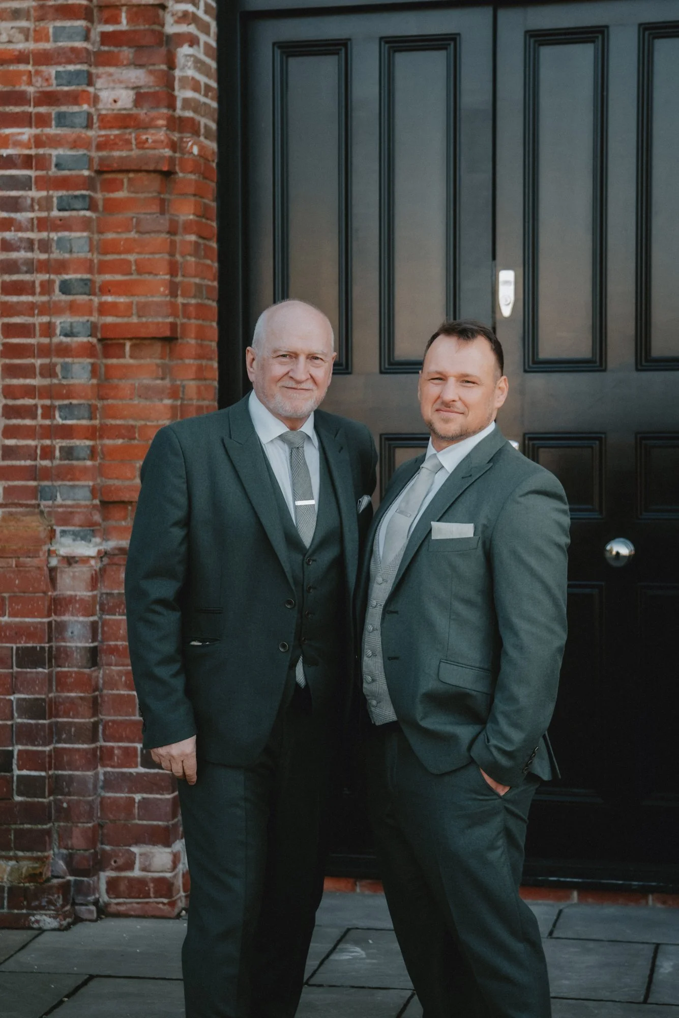 Two men in suits standing next to each other in front of a black door and a brick wall.