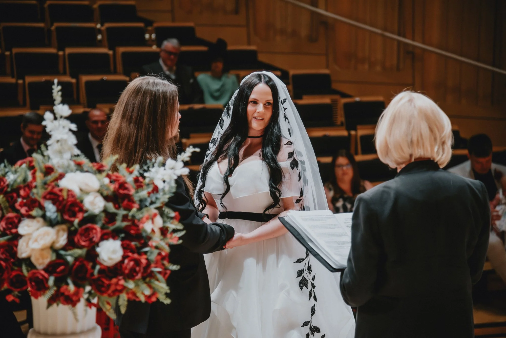 A bride and groom exchanging vows during a wedding ceremony inside a church, with a officiant reading from a book and floral arrangements nearby.