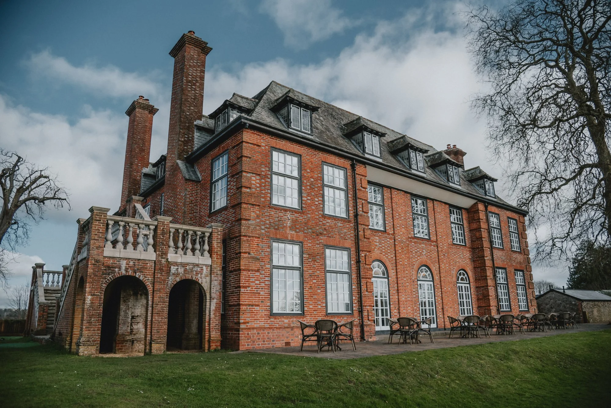 Large brick mansion with multiple windows and chimneys, outdoor patio with chairs, green lawn, leafless trees, partly cloudy sky in the background.