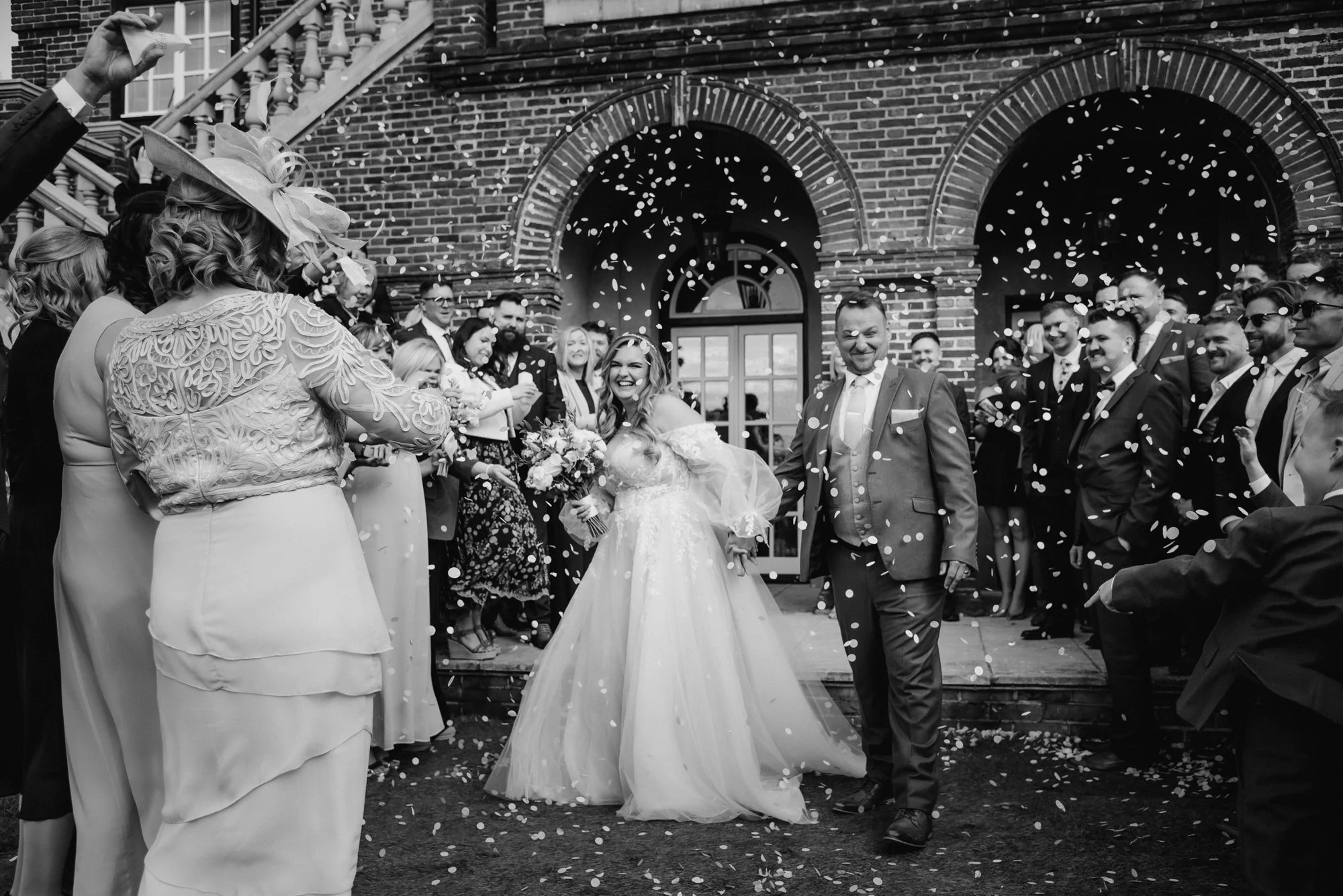 A black and white photo of a wedding celebration outside a brick building with a group of people throwing confetti, including a bride in a wedding gown holding a bouquet, and a groom in a suit smiling as they walk forward.