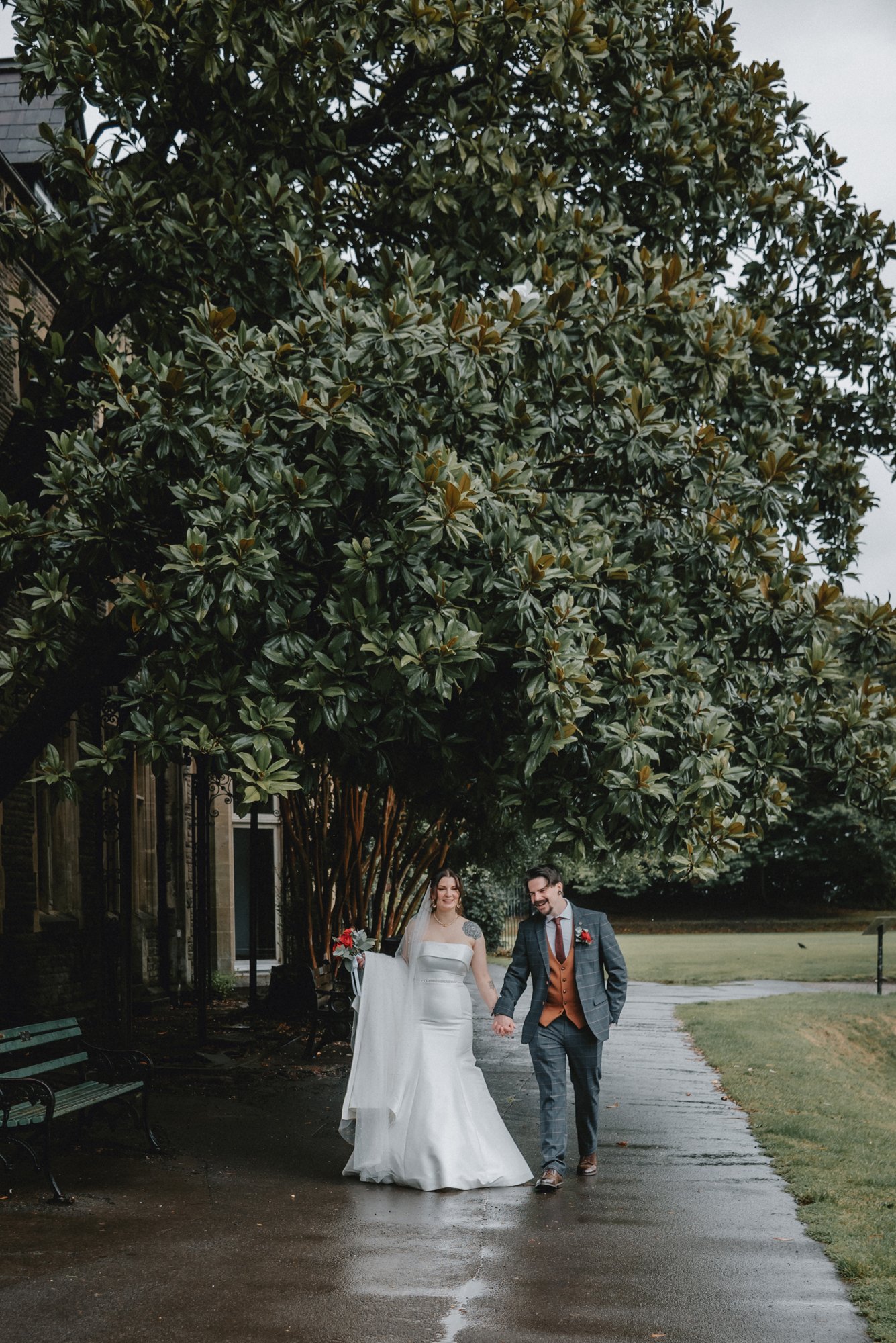 A newlywed couple walking hand in hand under a large leafy tree on a wet pathway, smiling and enjoying their wedding day.