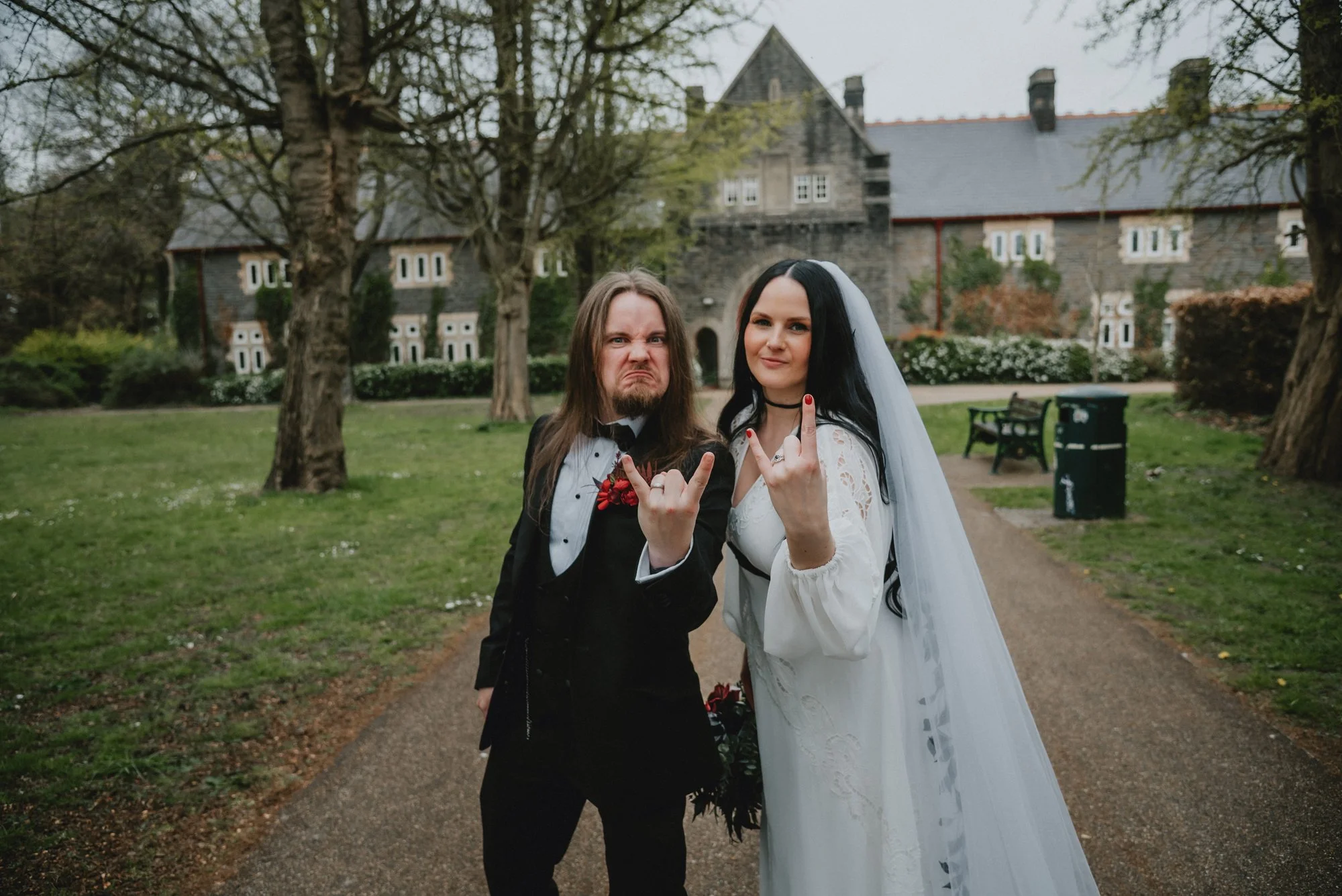 A couple in wedding attire making rebellious hand signs in a park with trees and a historic-looking building in the background.
