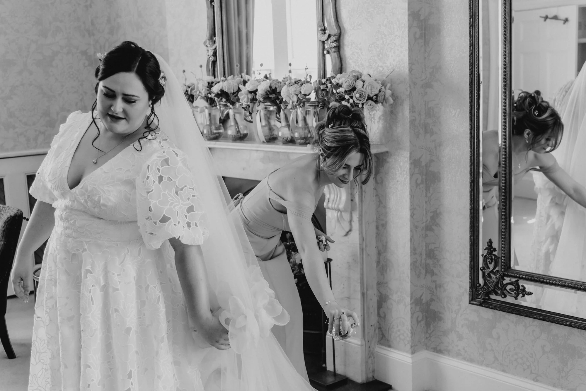 Two women in elegant dresses, one in a wedding gown, preparing for a wedding. One is holding the train of her gown while the other is adjusting her dress with a drink in hand. They are in a beautifully decorated room with a large mirror and floral ar