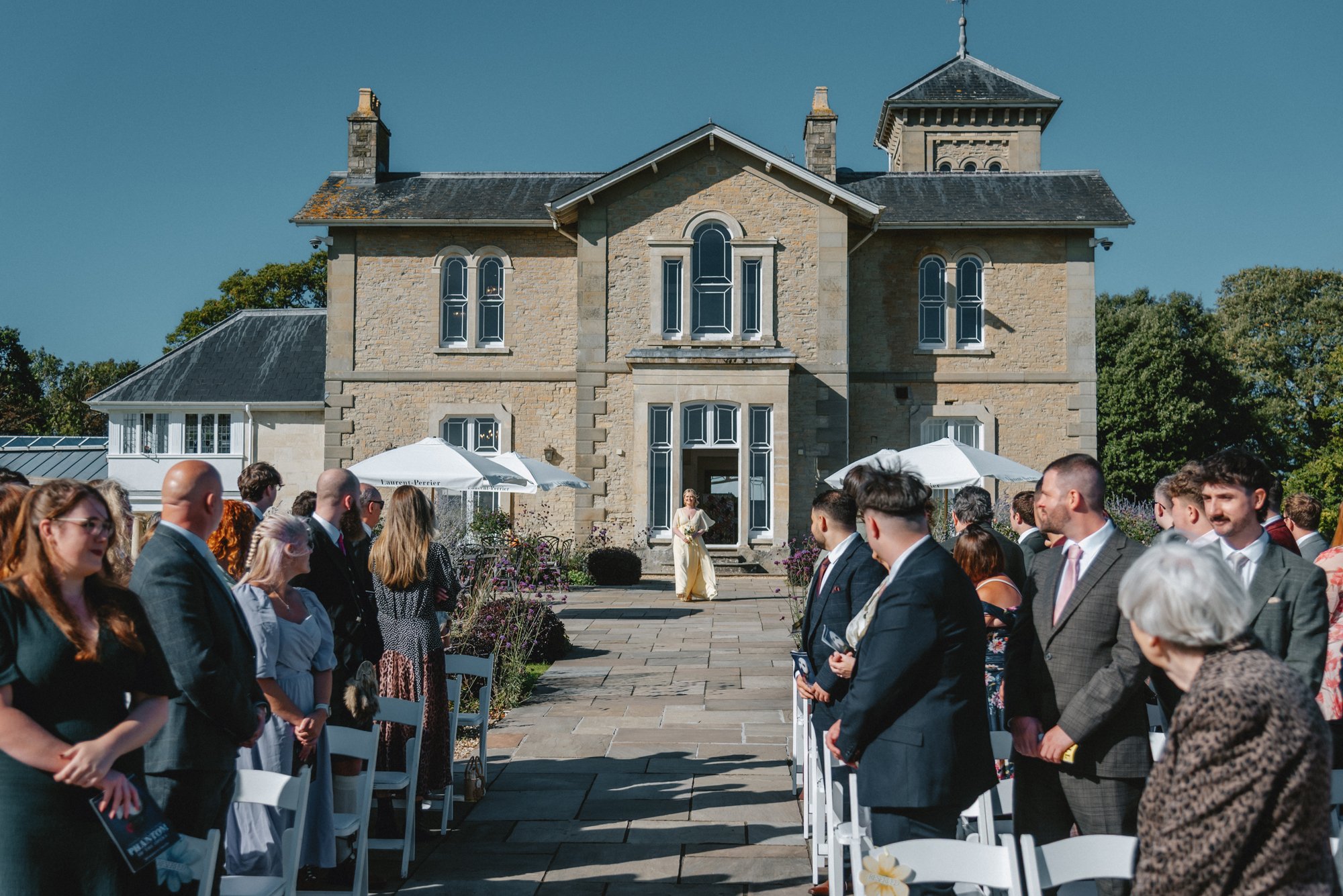 A wedding ceremony outdoors in front of a large stone house, with guests standing on either side of the aisle. The bride is walking toward the house doorway as the guests look on.
