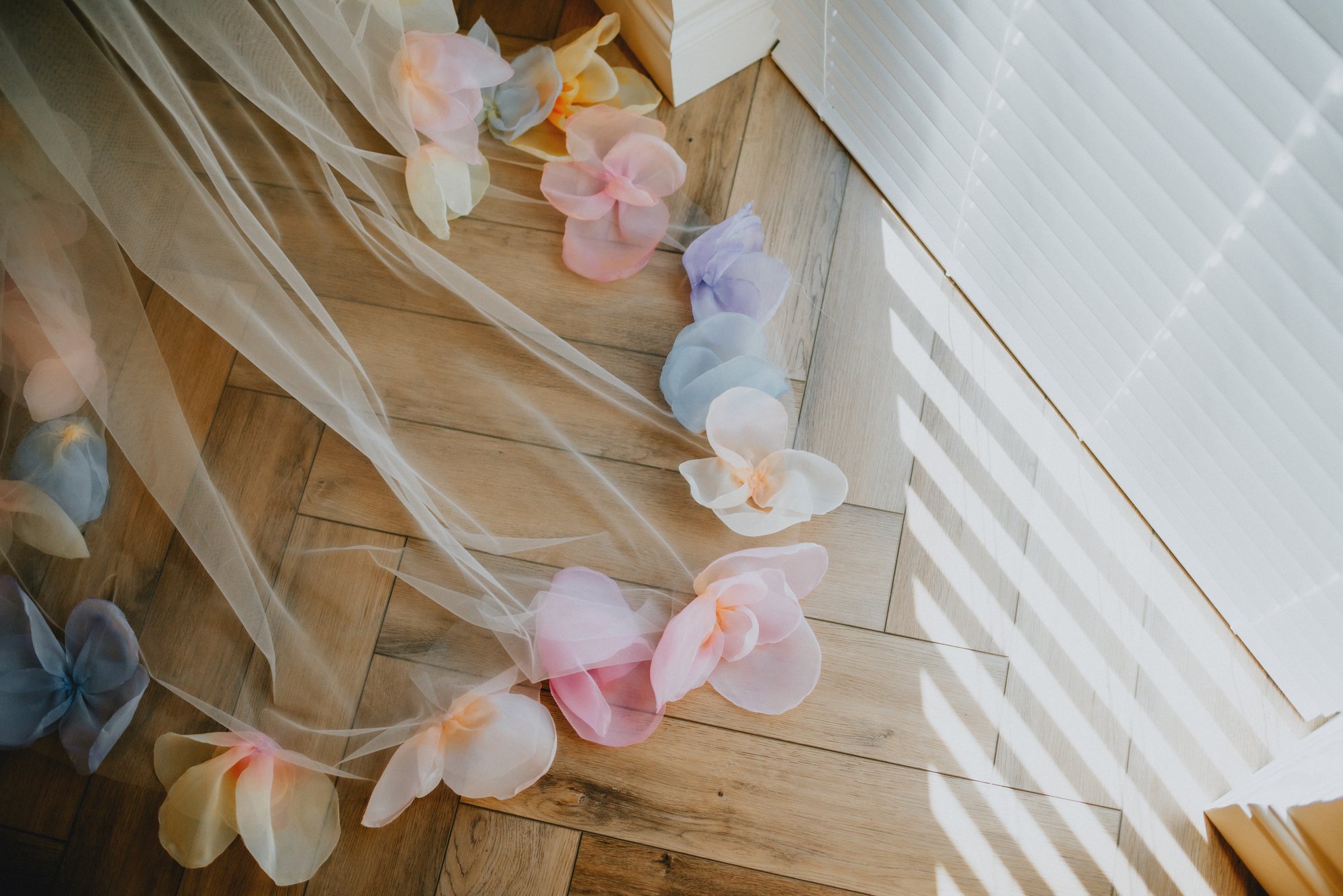 Colorful artificial flower petals scattered on a wooden floor next to a sheer curtain and window blinds with sunlight casting shadows.