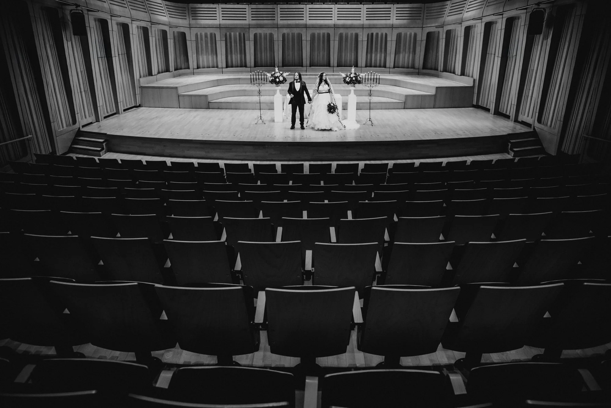 A black and white photo of a wedding ceremony on an empty stage in an auditorium. The couple stands in the center, holding hands, with flowers and candles as decorations.