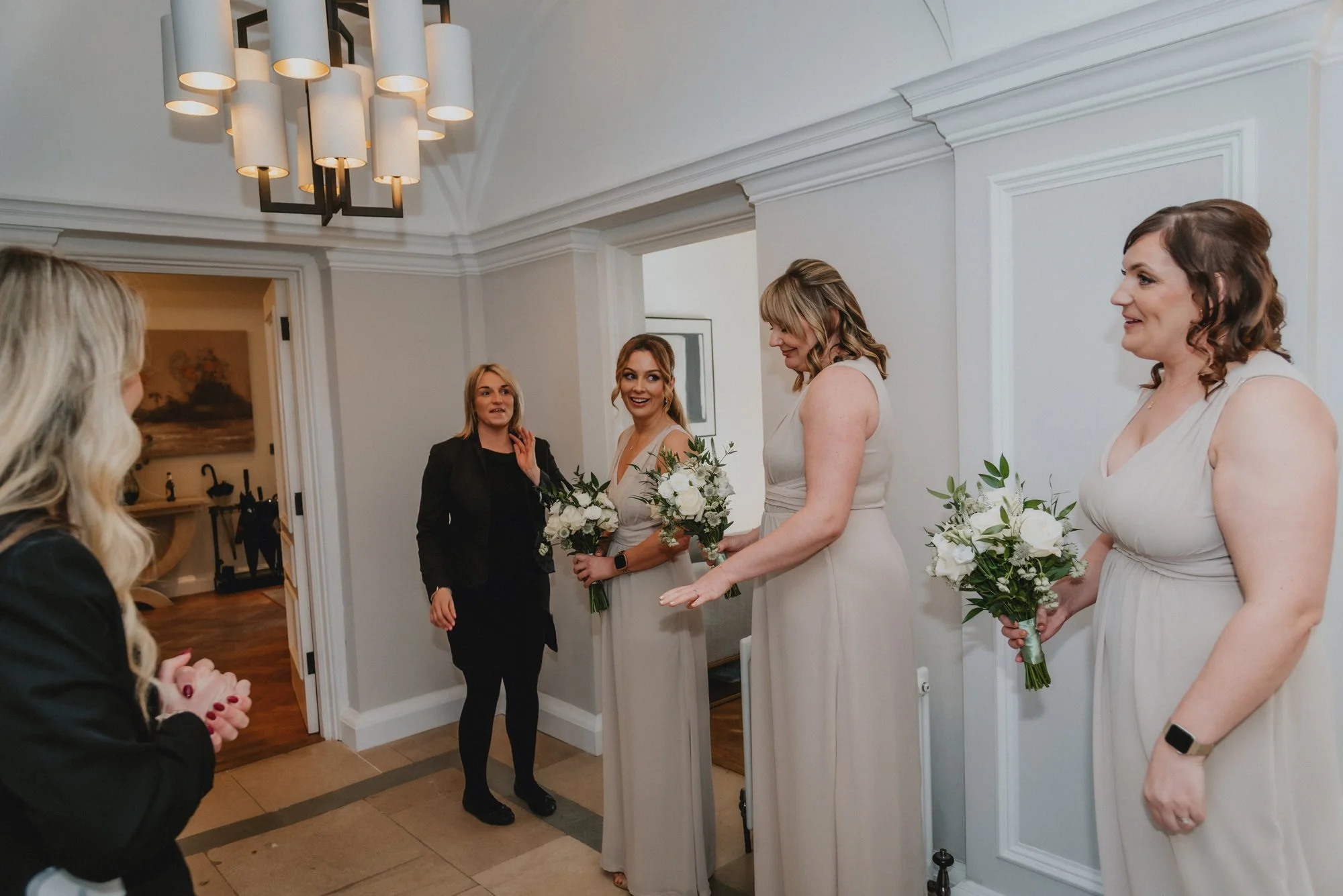 A bride with three bridesmaids in a room, all holding bouquets of white flowers. They are smiling and engaging in conversation with a woman in black.