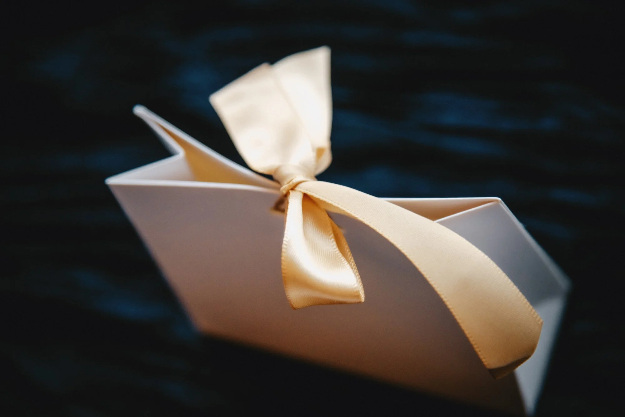 A white gift box with a cream-colored satin ribbon tied in a bow, placed on a dark, textured surface.