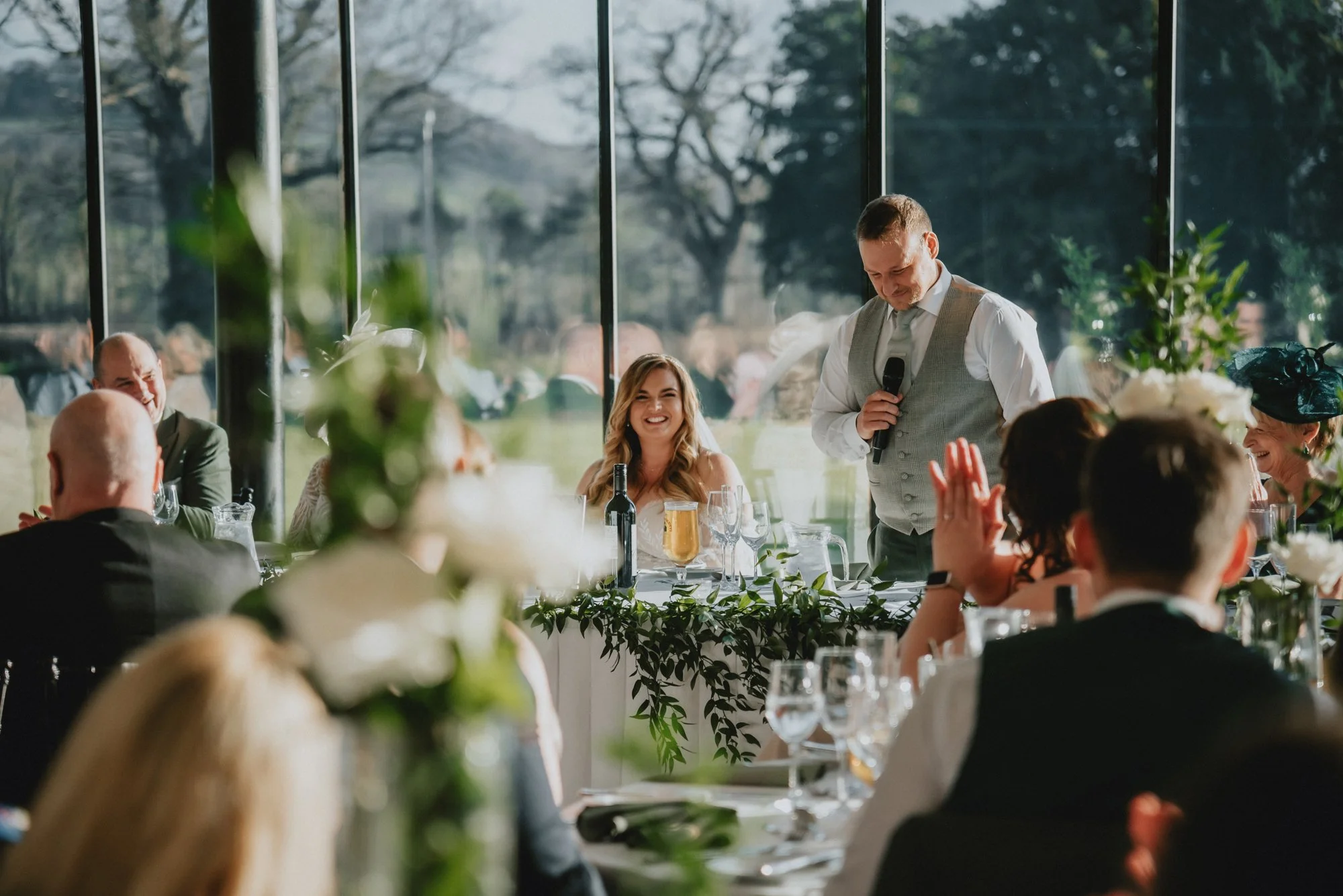 A man giving a speech at a wedding reception, with guests seated at tables decorated with flowers and greenery, smiling and clapping.