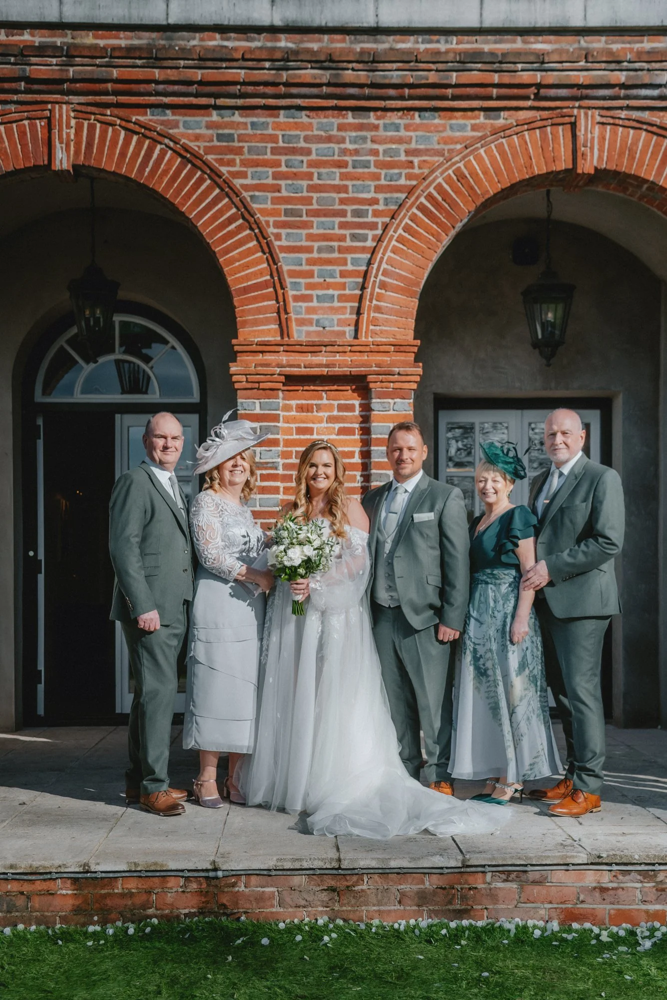 A group of six people standing in front of a brick building with arches, dressed in formal wedding attire, with a bride holding a bouquet of white flowers in the center.