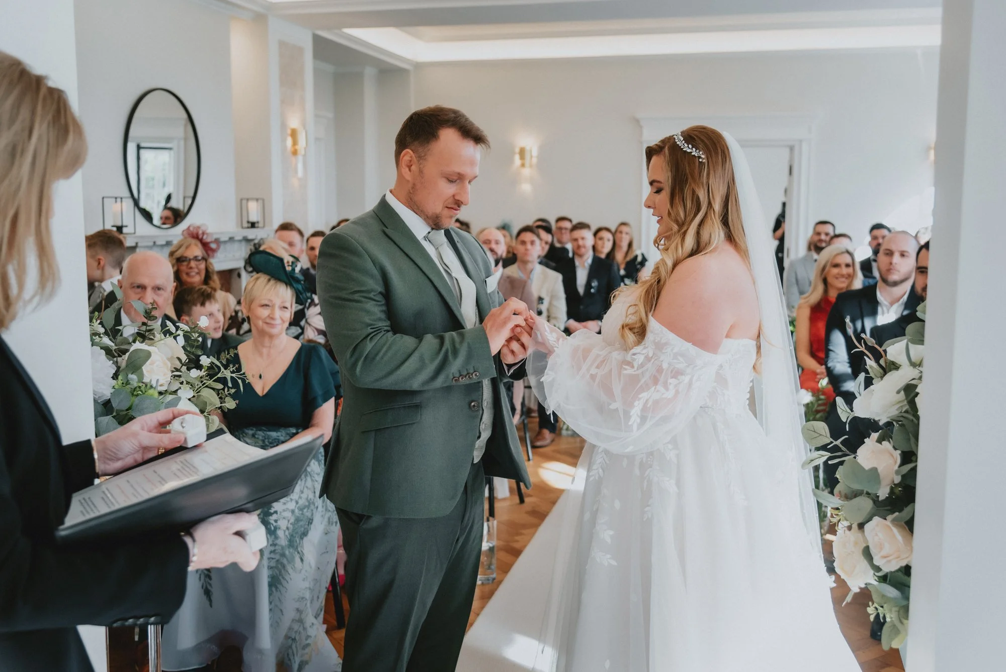 A bride and groom exchanging wedding vows during their ceremony in a bright, elegant room, surrounded by seated guests and floral decorations.