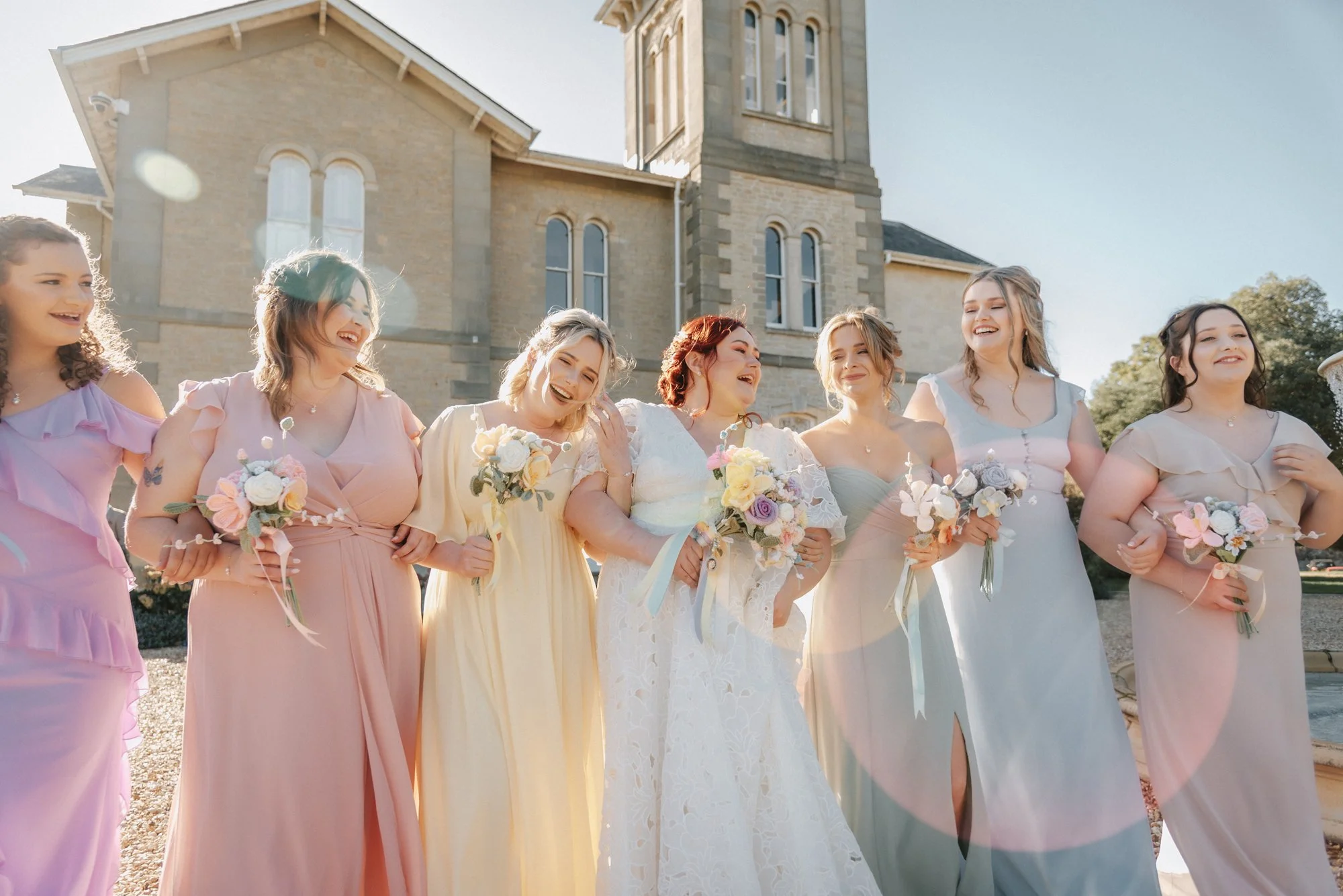 Group of women in dresses smiling and holding bouquets during a wedding celebration outside a church.