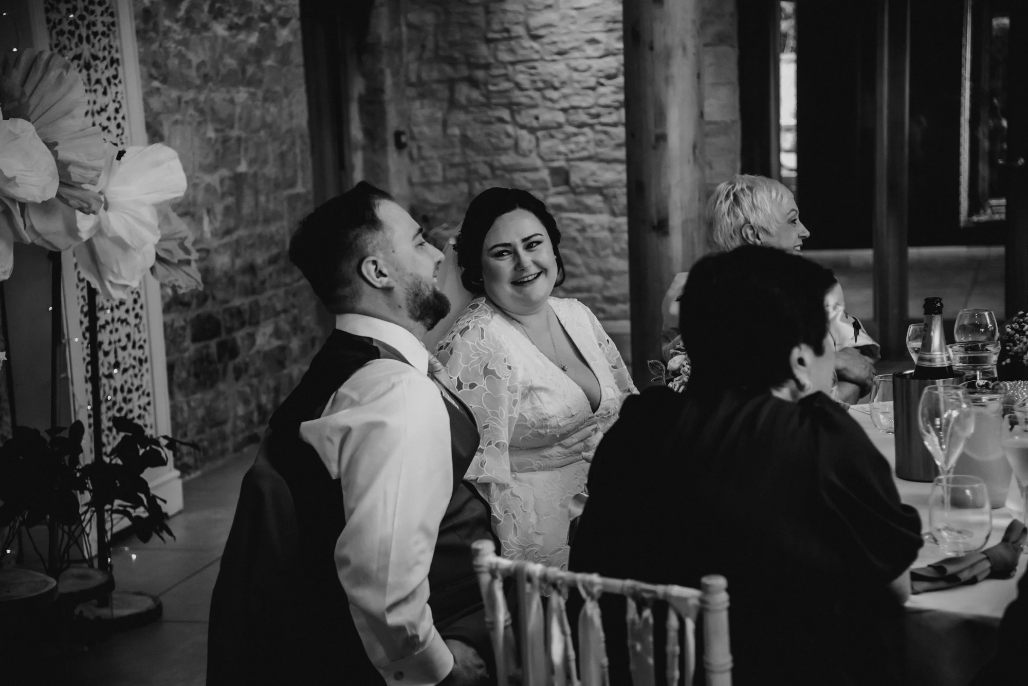 People sitting at a wedding reception table, with one woman smiling and looking at the camera, and others talking and laughing, in a rustic venue with stone walls and wooden beams.