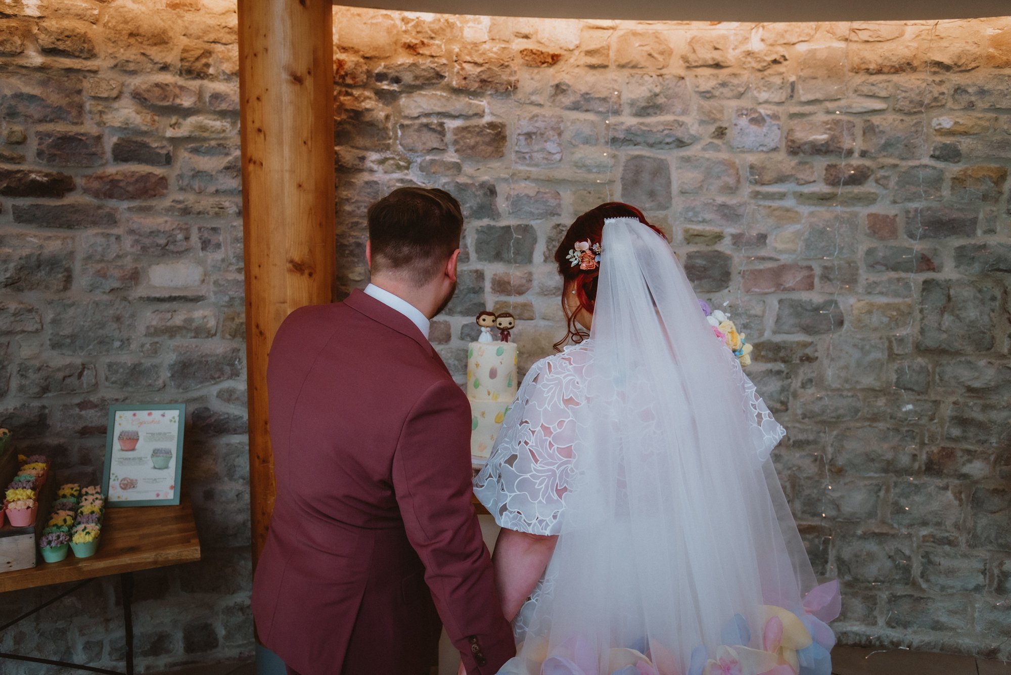A bride and groom standing together in front of a stone wall, holding hands and facing a wedding cake with figurines on top.