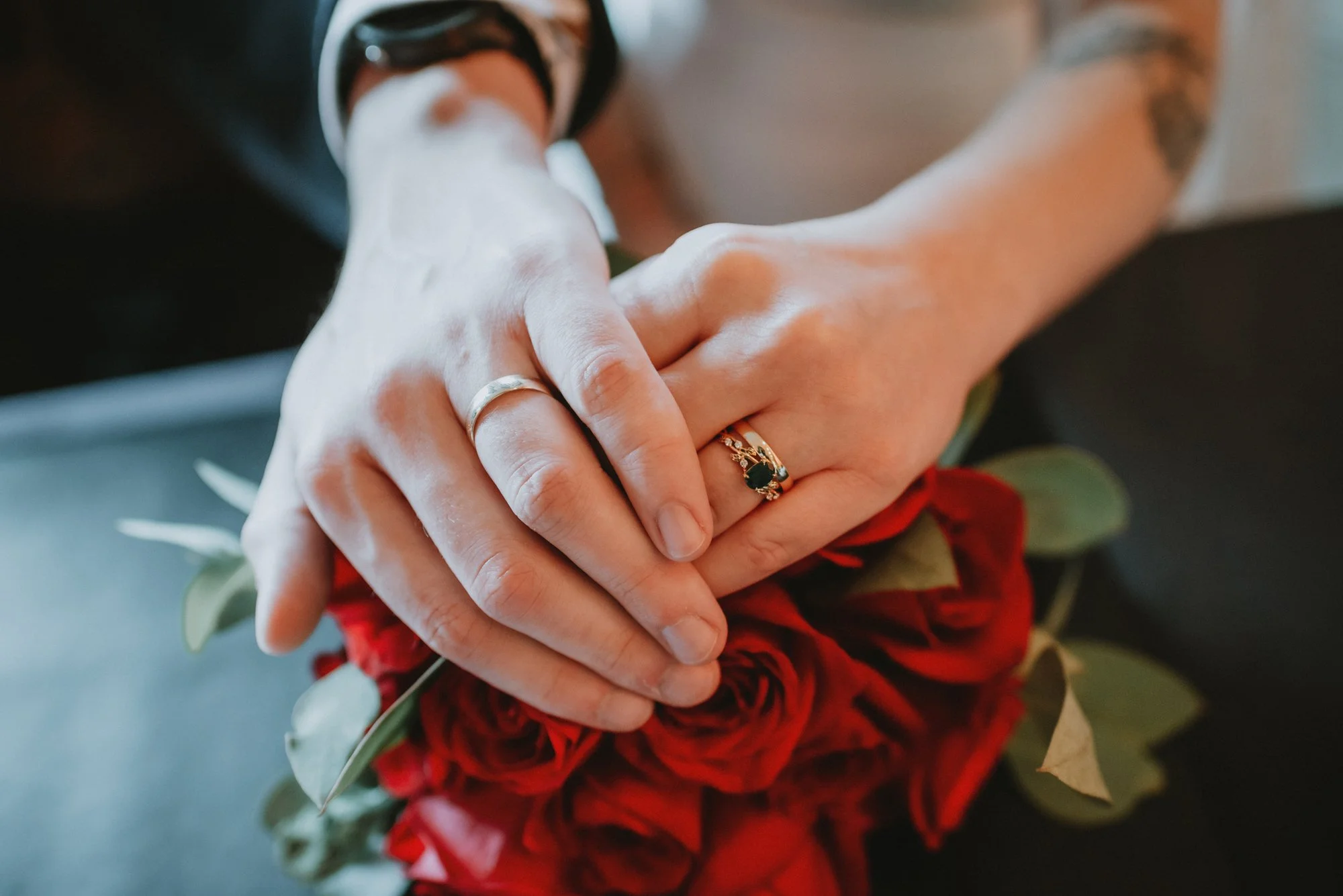 Couple's hands with wedding rings, holding a bouquet of red roses and green leaves.