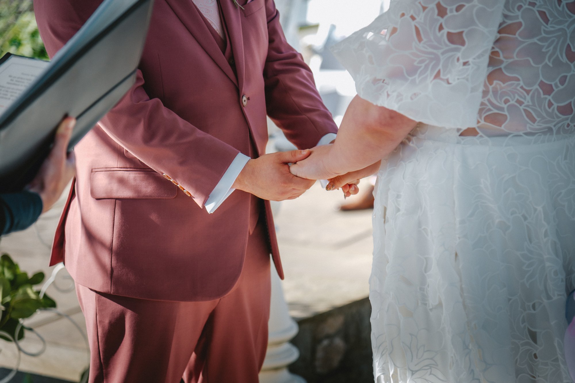 Close-up of a couple holding hands during a wedding ceremony. The groom is wearing a maroon suit, and the bride is in a white lace wedding dress. An officiant is holding a book or tablet on the left side of the image.
