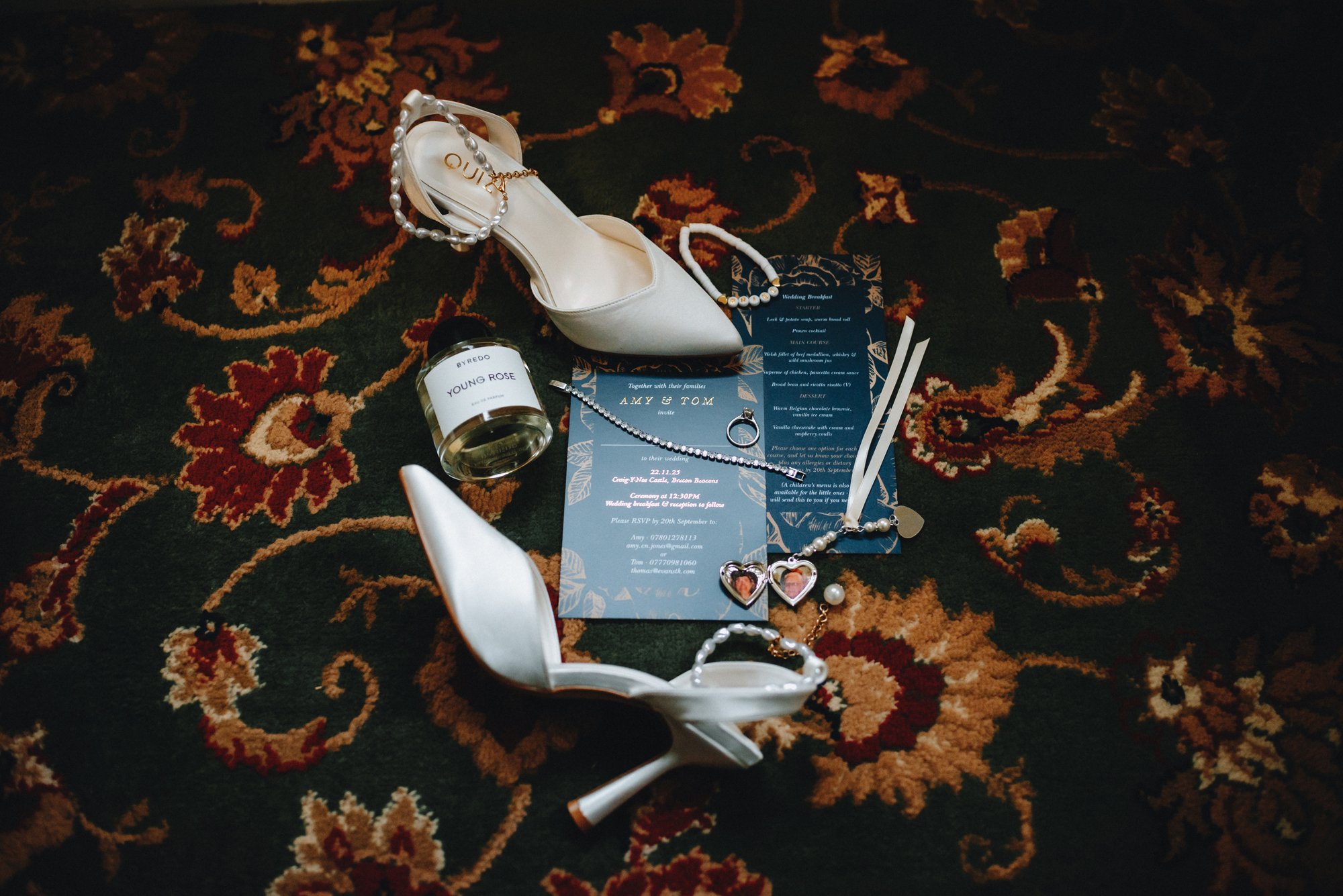 Wedding accessories including white high-heeled shoes, pearl jewelry, a perfume bottle labeled 'Young Rose,' a wedding invitation, and a bottle of perfume on a patterned carpet.