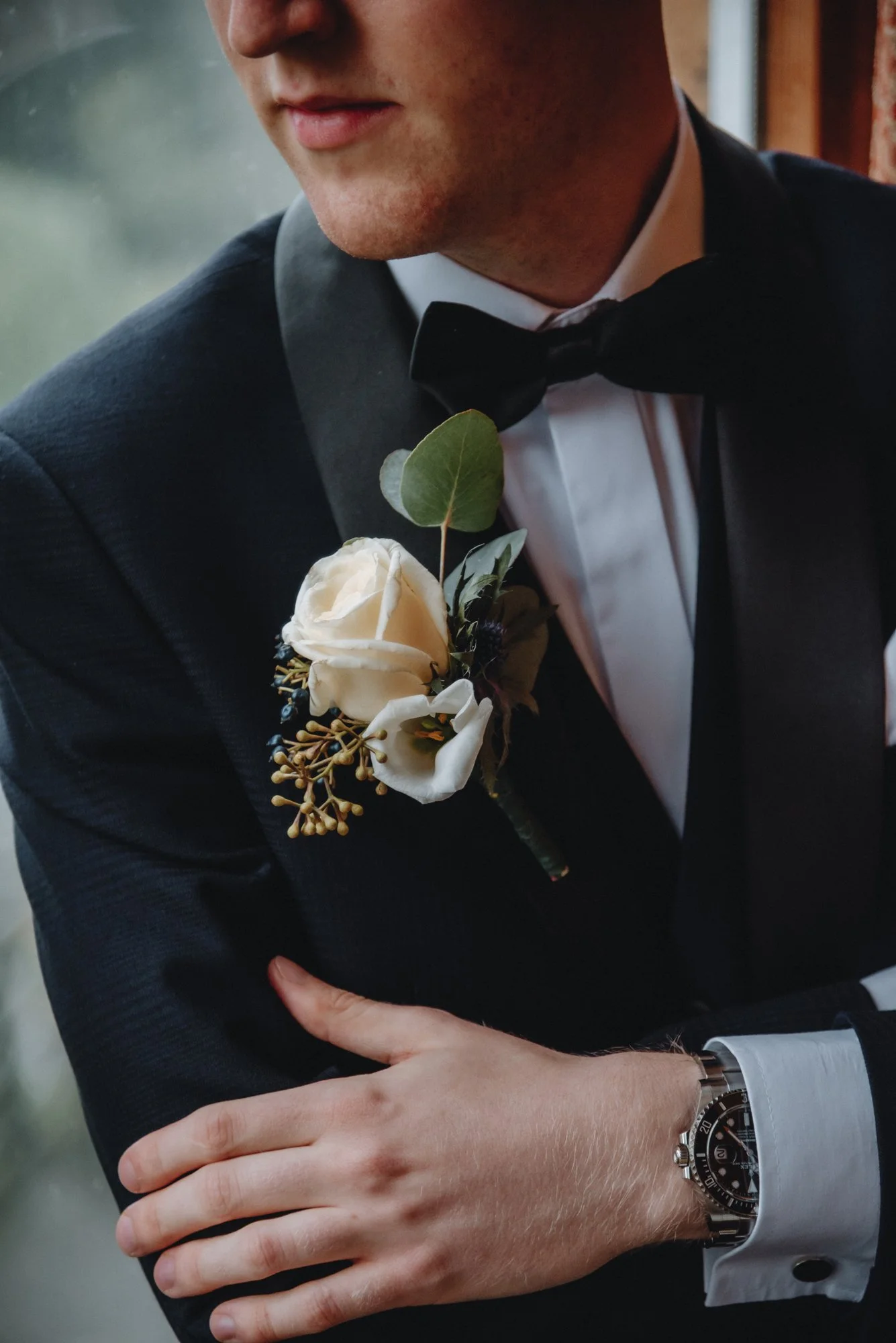 A man dressed in formal black tuxedo with a white shirt and bow tie, wearing a wristwatch, has a white rose boutonniere with greenery on his left lapel.