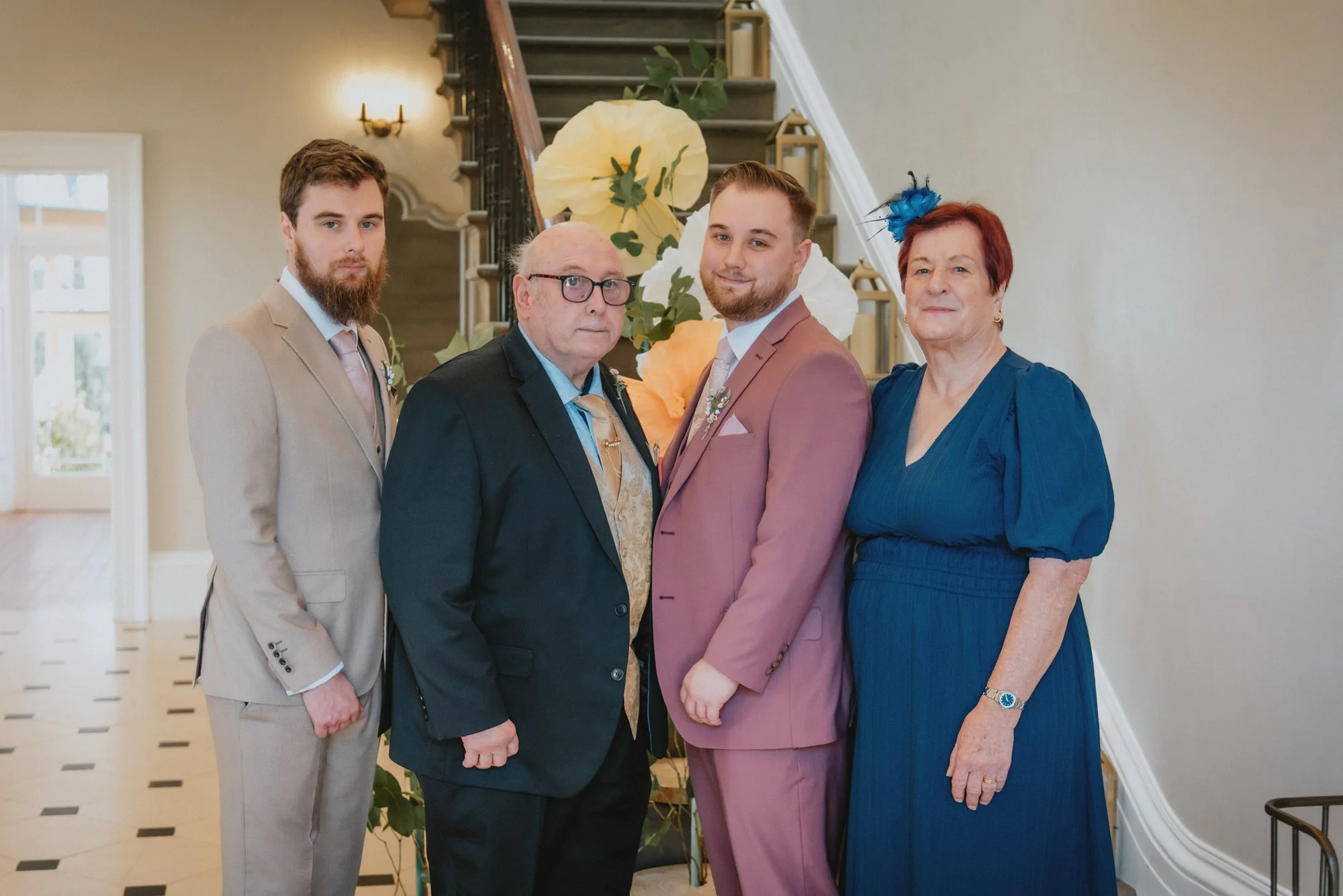 Four people dressed in formal attire standing in front of a staircase decorated with large paper flowers, in an elegant interior.